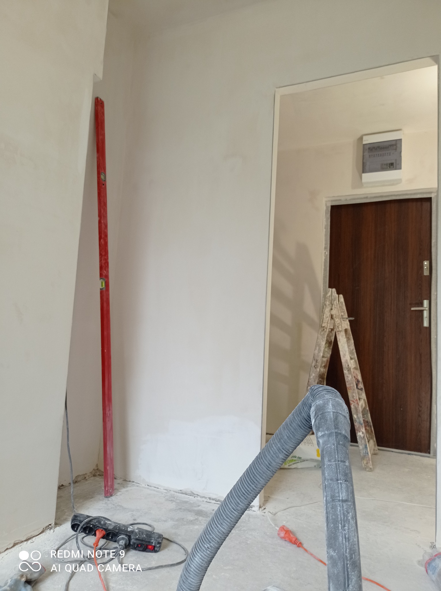 Wnętrze w trakcie prac wykończeniowych: poziomica przy ścianie, widoczny wąż odkurzacza przemysłowego i drabina. Ściany przygotowane pod malowanie.