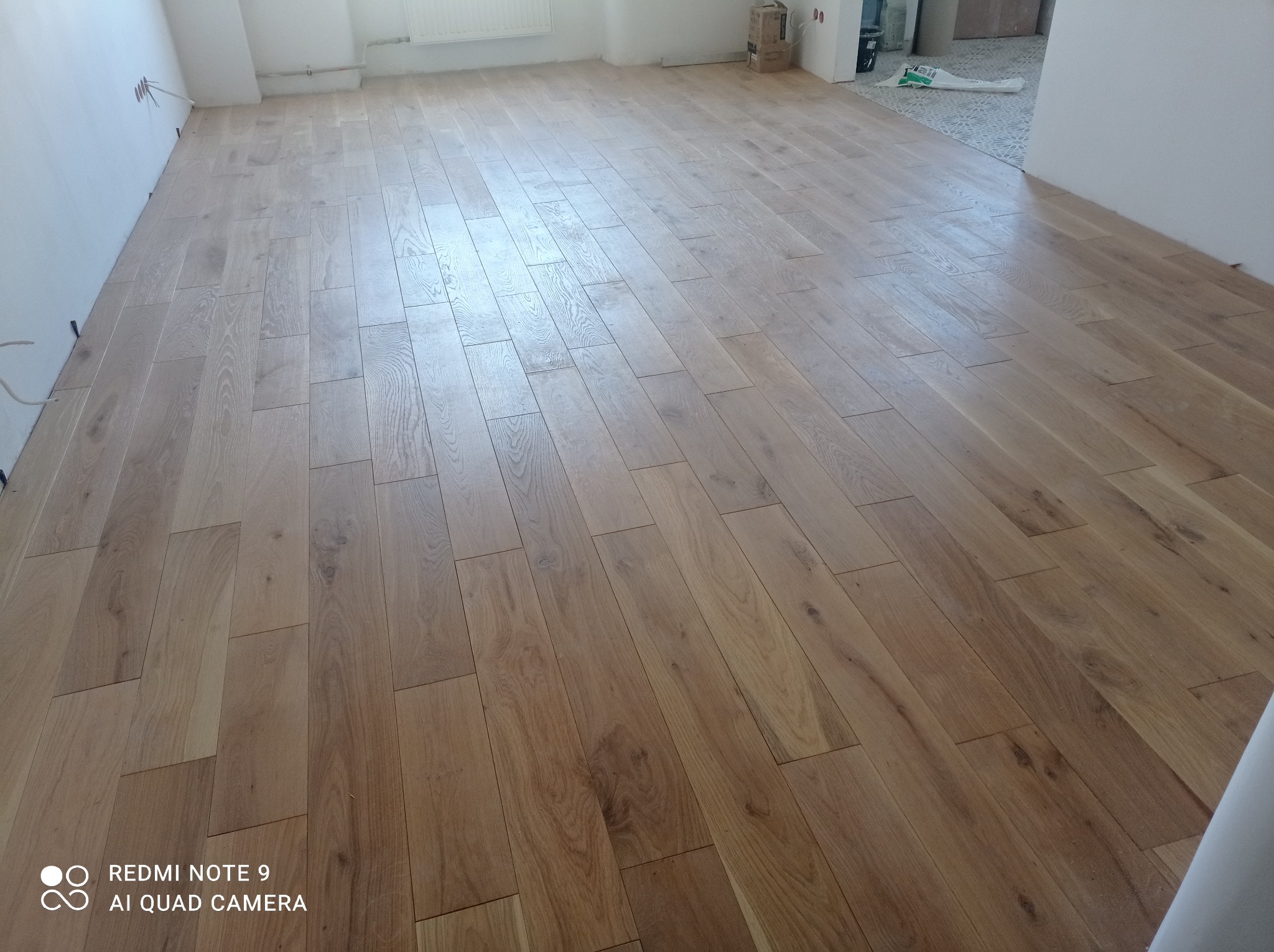 Ułożona podłoga z paneli imitujących drewno w jasnym odcieniu, widok z góry, fragment ściany i podłogi w innym pomieszczeniu w tle.