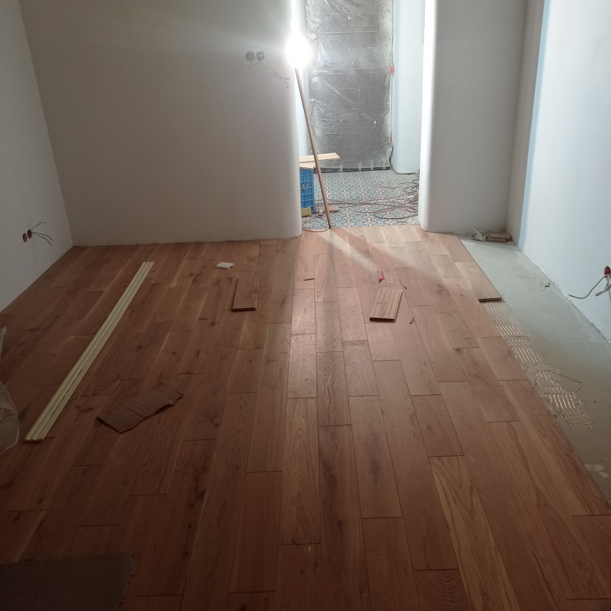 Układanie parkietu w pokoju z widocznymi fragmentami podłogi, listwami przypodłogowymi i narzędziami. W tle zabezpieczone folią drzwi.