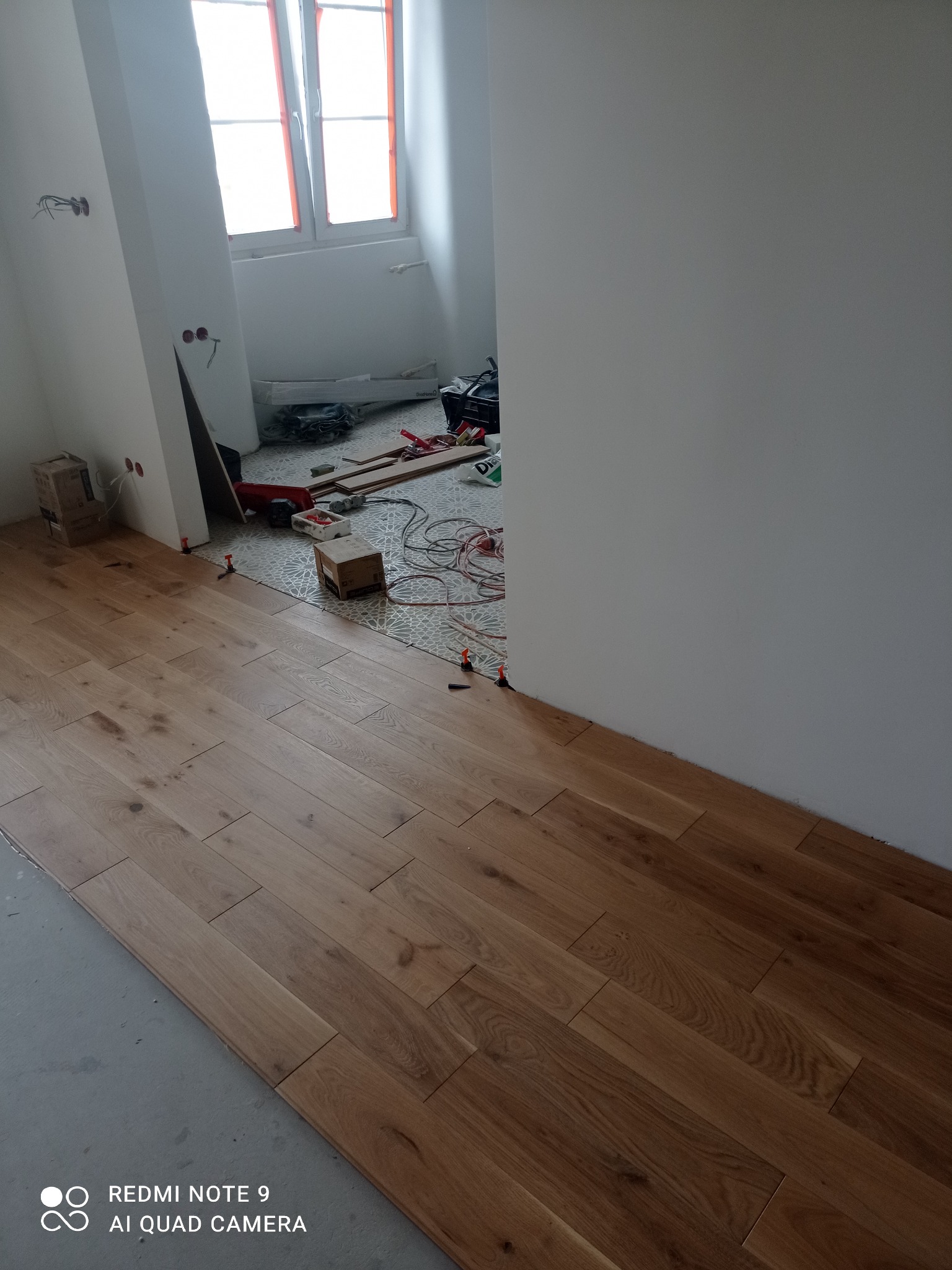 Układanie drewnianej podłogi w jodełkę francuską. Widoczne płytki podłogowe w sąsiednim pomieszczeniu i narzędzia pracy. Użycie poziomowania płytek.