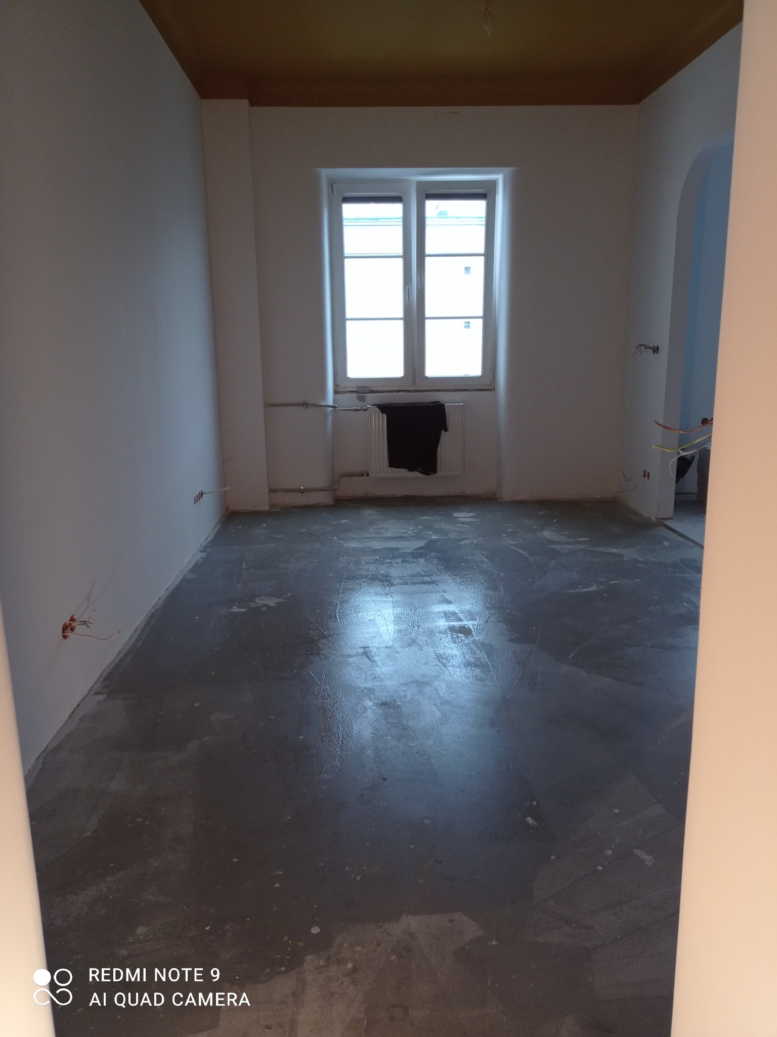 Szara, mokra wylewka anhydrytowa w pustym pokoju z oknem. Widoczne wystające kable elektryczne ze ściany. Przy oknie wisi czarny materiał.