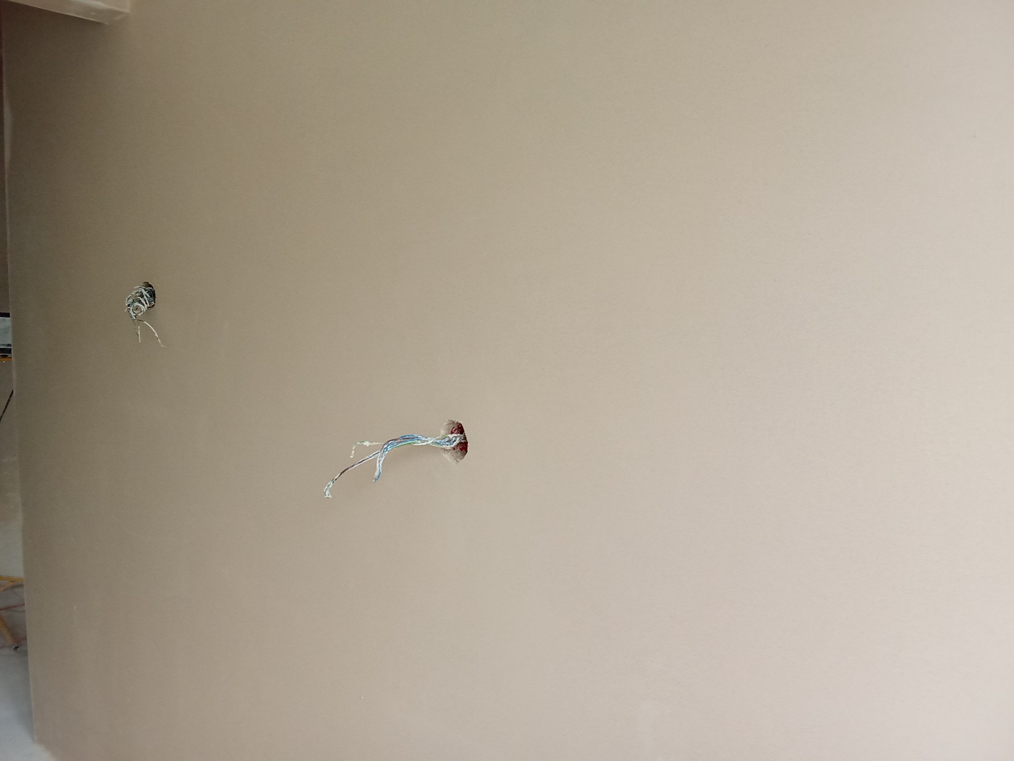 Gładka, beżowa ściana po szpachlowaniu z dwoma otworami na kable elektryczne. Widoczne przewody w izolacji. Przygotowanie do dalszych prac wykończeniowych.