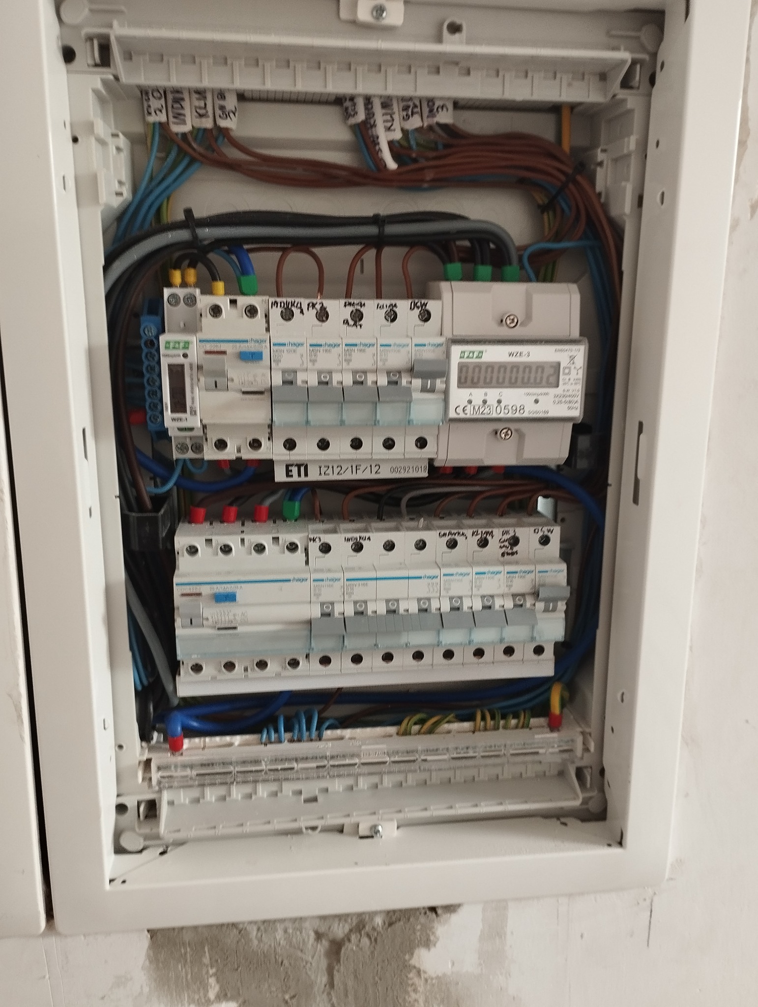 Otwarta skrzynka elektryczna z widocznymi bezpiecznikami, przewodami o różnych kolorach i licznikiem energii elektrycznej wewnątrz, zamontowana na surowej ścianie.