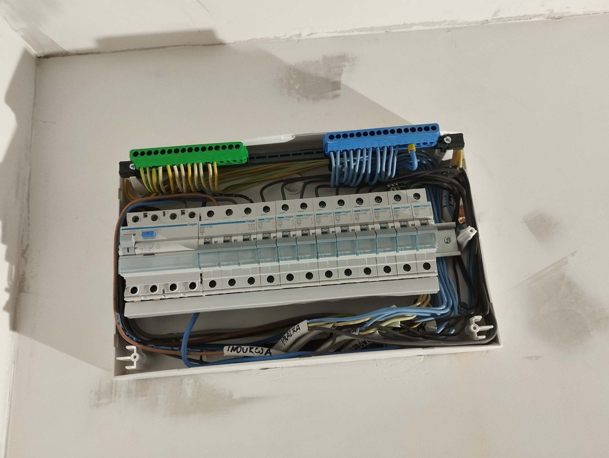 Otwarta skrzynka rozdzielcza z zainstalowanymi bezpiecznikami, widoczne przewody z oznaczeniami 'Inoukcją' i 'Pralka', listwy zaciskowe zielone i niebieskie, montaż na surowej ścianie.