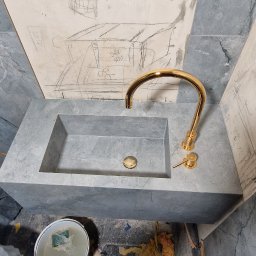 Funt BUD - Szara umywalka z baterią w kolorze złotym, na ścianie rysunki ołówkiem. Widoczne ślady prac remontowych.