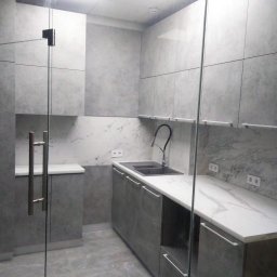 Funt BUD - Nowoczesna, szara kuchnia z marmurowym blatem i szklanymi drzwiami. Minimalistyczny design, oświetlenie sufitowe, widoczne gniazdka elektryczne.