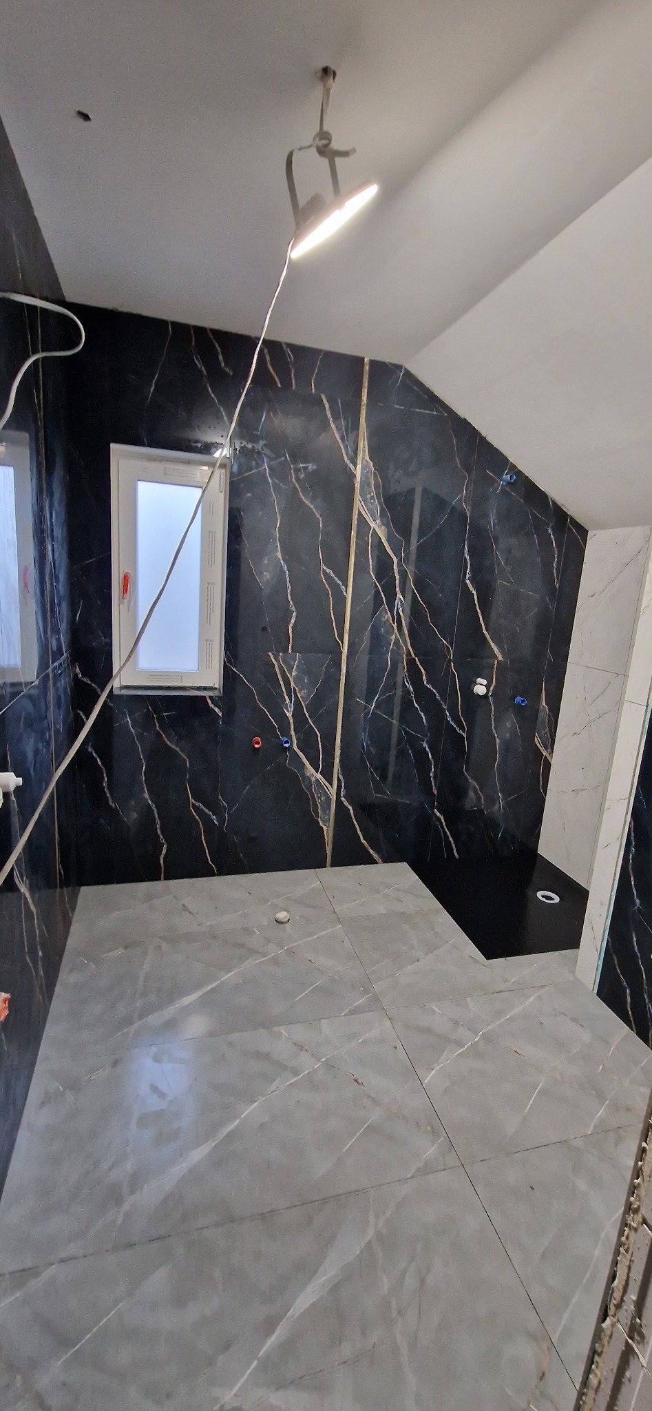 Wykończona łazienka na poddaszu z czarnymi i szarymi płytkami imitującymi marmur, oświetlona lampą budowlaną. Widoczny brodzik i okno.