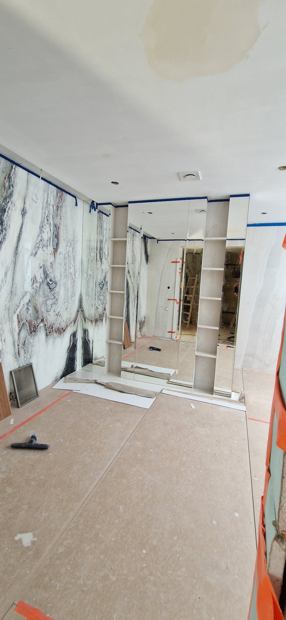 Wnętrze pokoju z marmurowymi ścianami i lustrami w trakcie wykończenia. Widoczne narzędzia i zabezpieczenia. Podłoga częściowo wyłożona panelami.