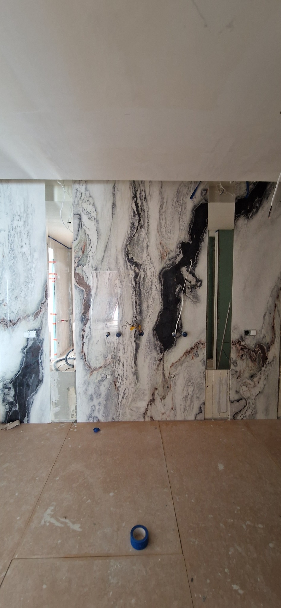 Montaż marmurowych płyt ściennych w łazience, widoczne puszki elektryczne i taśma malarska na podłodze. Fragment lustra wkomponowany w ścianę.