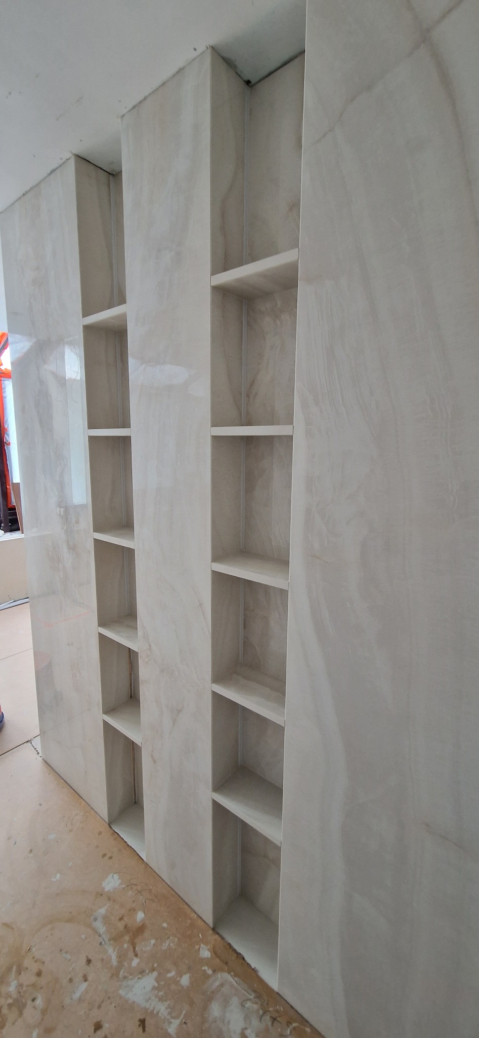 Wykończona ściana z wnękami wyłożona płytkami imitującymi marmur. Widoczne półki wnękowe. Podłoga w trakcie prac wykończeniowych.