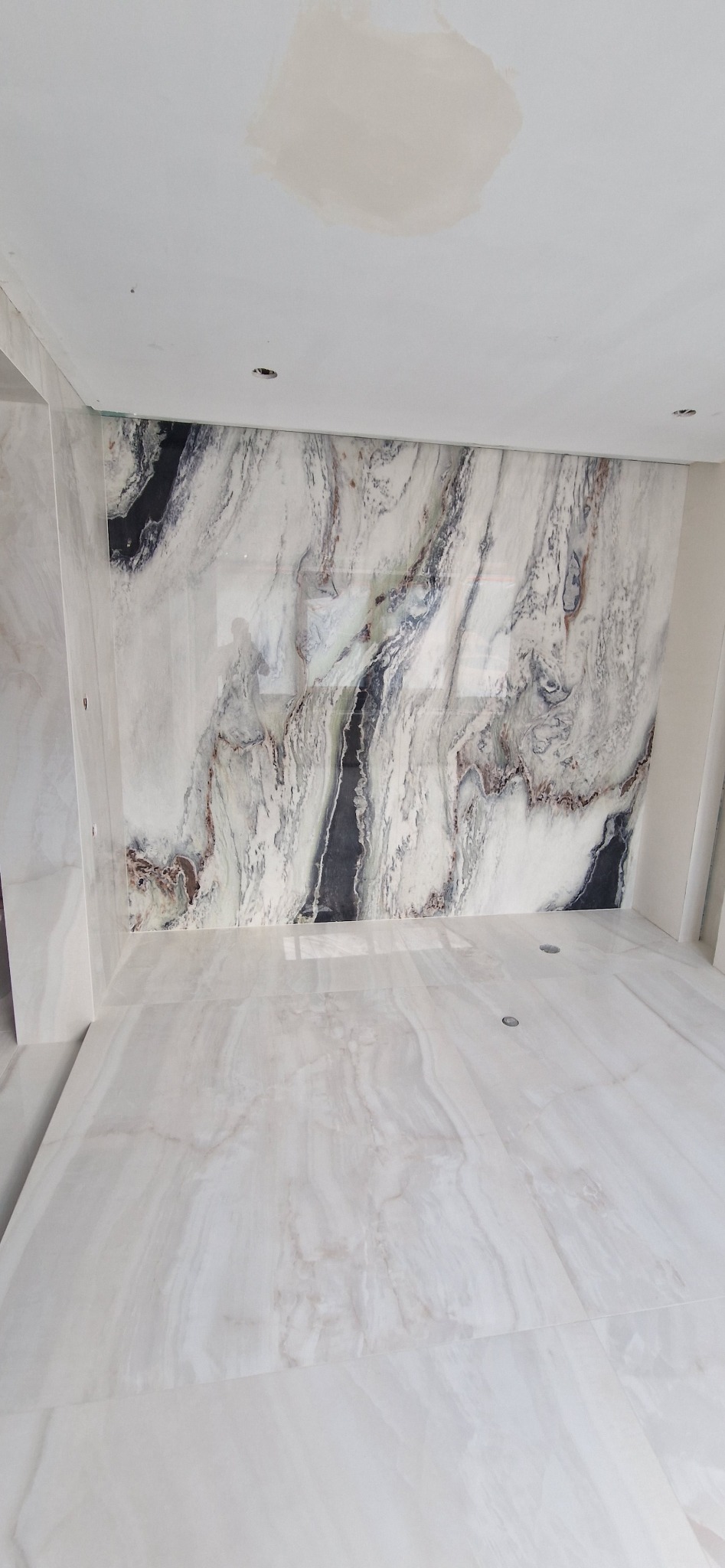 Wykończona łazienka z marmurowymi płytami na ścianach i podłodze. Na suficie widoczny fragment świeżo położonej gładzi. Minimalistyczny design.