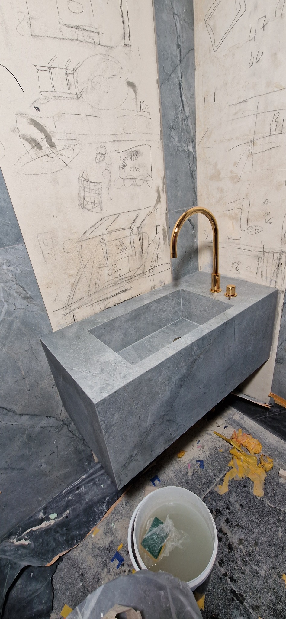 Szara, kamienna umywalka z złotą armaturą na tle ściany z rysunkami, w trakcie prac wykończeniowych. Widoczne narzędzia i resztki materiałów.