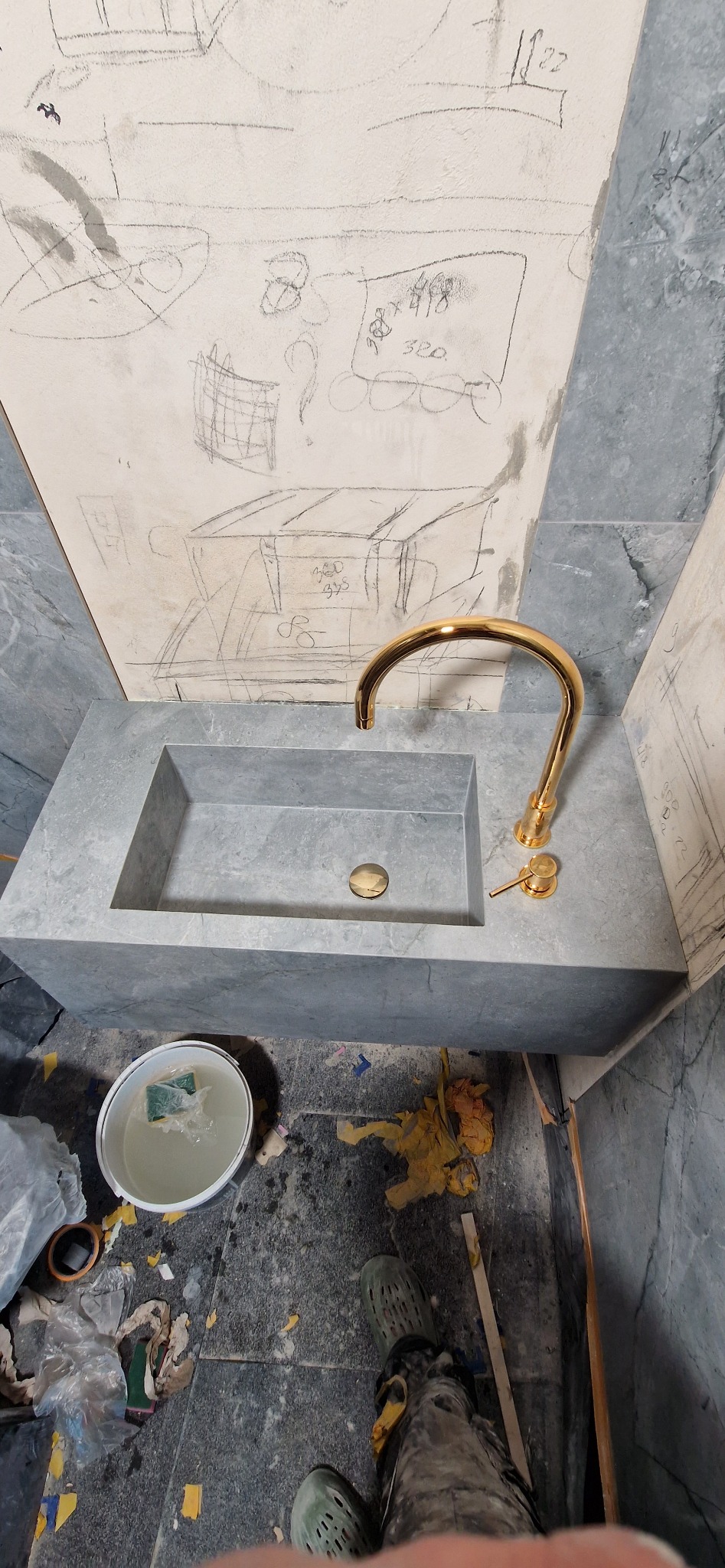 Szara umywalka z baterią w kolorze złotym, na ścianie rysunki ołówkiem. Widoczne ślady prac remontowych.