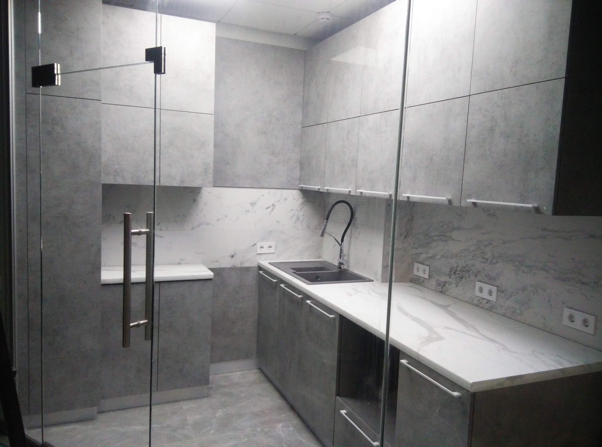 Nowoczesna, szara kuchnia z marmurowym blatem i szklanymi drzwiami. Minimalistyczny design, oświetlenie sufitowe, widoczne gniazdka elektryczne.