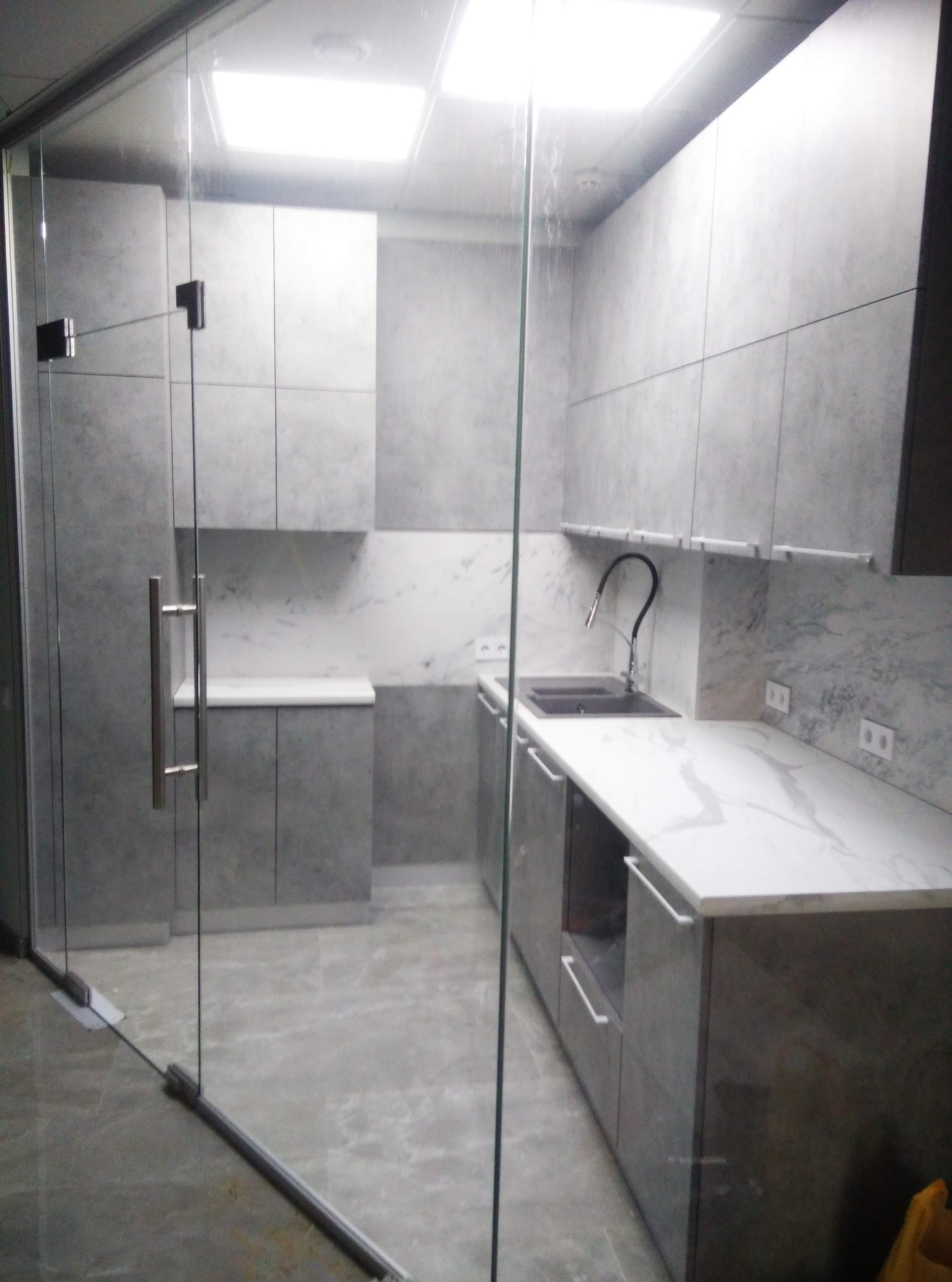 Nowoczesna łazienka z szarą zabudową, marmurowym blatem i płytkami imitującymi beton. Całość widoczna przez szklane drzwi.