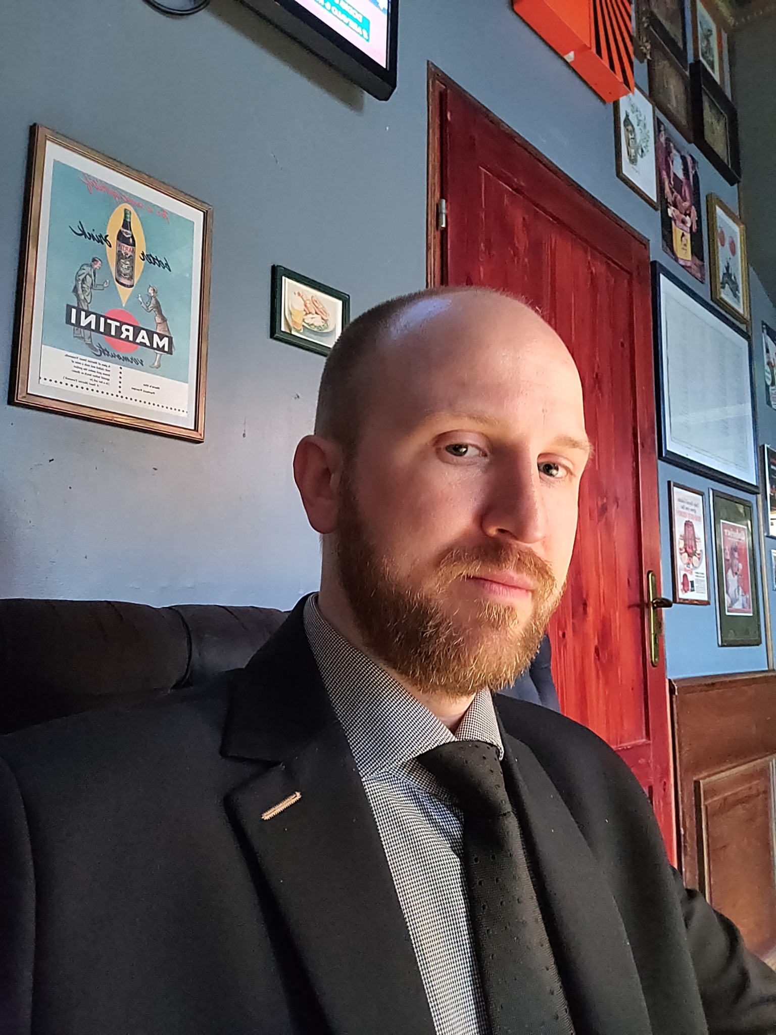 Portret mężczyzny z rudą brodą w garniturze, siedzącego na tle ściany z plakatami i czerwoną framugą drzwi. Profesjonalny wizerunek.