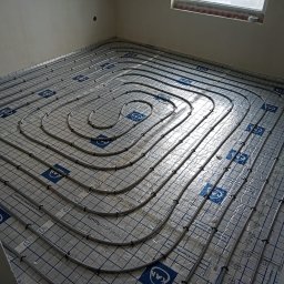 INSTALATORSTWO SANITARNE I GRZEWCZE RAFAŁ KOŁECKI - Rozłożone rury ogrzewania podłogowego na folii z nadrukiem siatki i logo KAN, widoczne uchwyty mocujące rury, surowy stan wykończenia wnętrza.