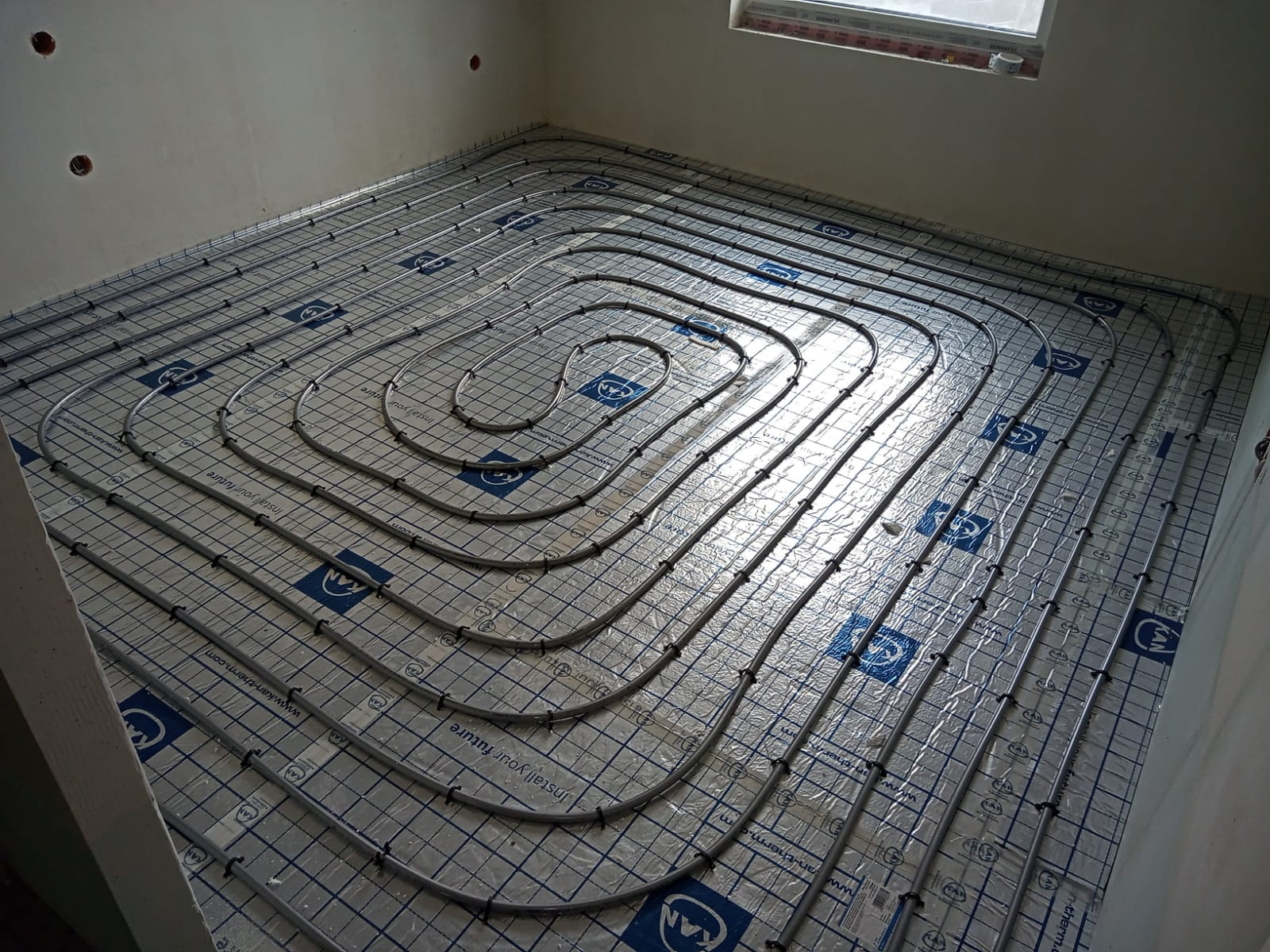 Rozłożone rury ogrzewania podłogowego na folii z nadrukiem siatki i logo KAN, widoczne uchwyty mocujące rury, surowy stan wykończenia wnętrza.