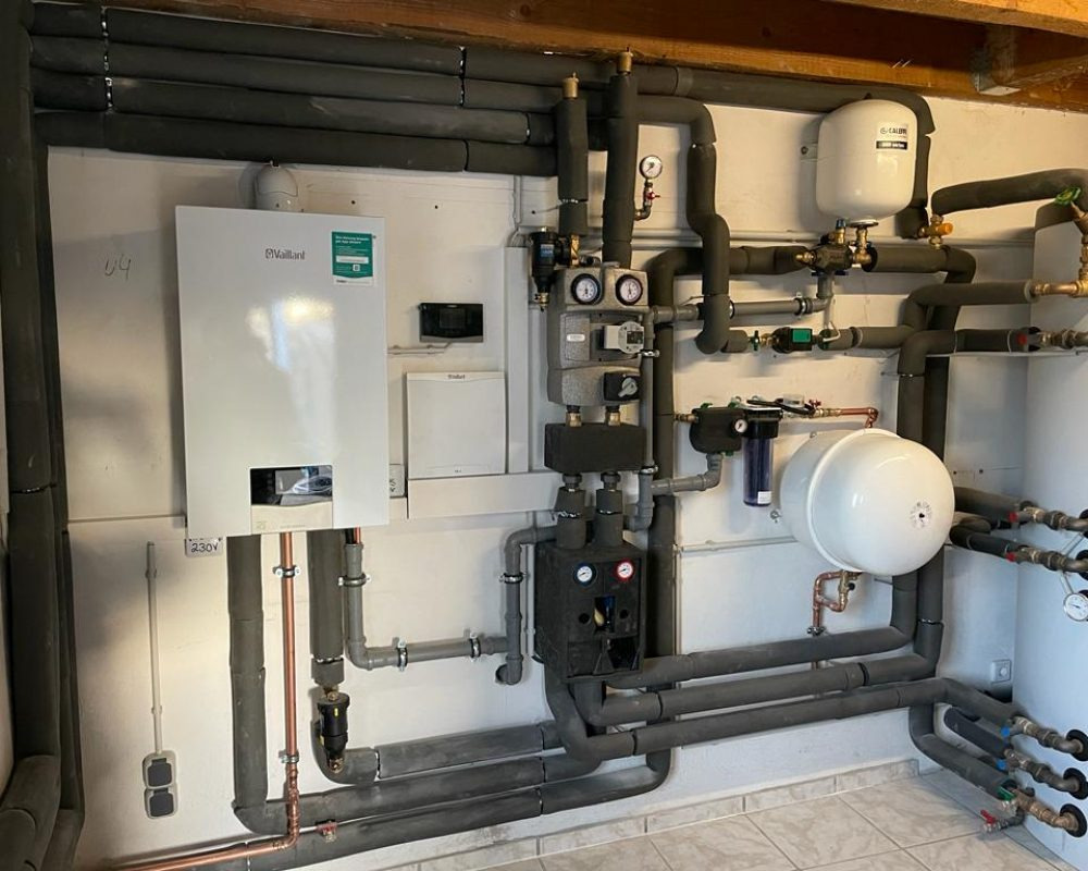 Złożona instalacja centralnego ogrzewania z kotłem gazowym, zbiornikami wyrównawczymi i rurami izolowanymi termicznie, zamontowana na ścianie w pomieszczeniu technicznym z wykończeniem w jasnych...