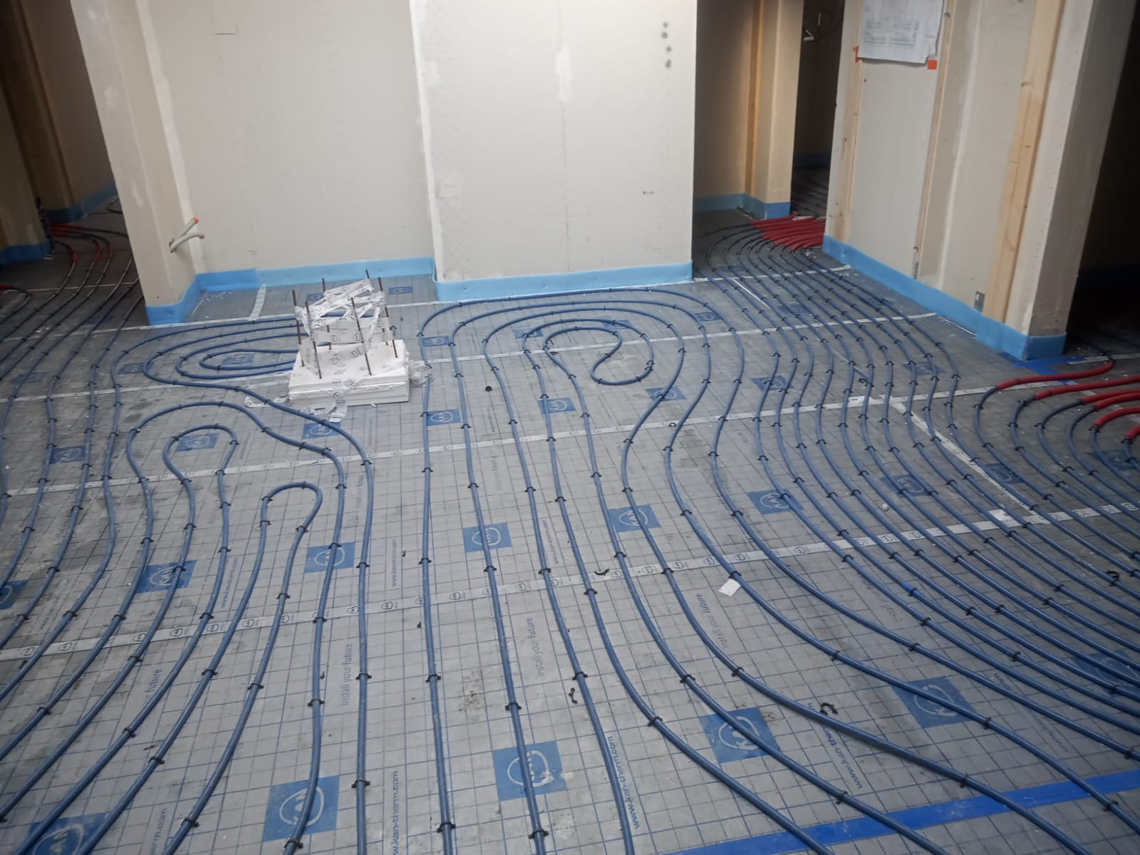 Montaż ogrzewania podłogowego: niebieskie rury ułożone w pętle na szarej macie izolacyjnej z niebieskimi brzegami, widoczne mocowania i fragmenty czerwonych rur, surowy stan wykończenia wnętrza.