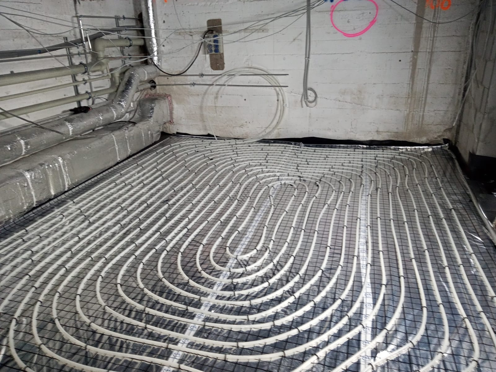 Montaż ogrzewania podłogowego: białe rurki ułożone w pętle na czarnej siatce, widoczne przewody instalacyjne przy suficie w surowym wnętrzu.
