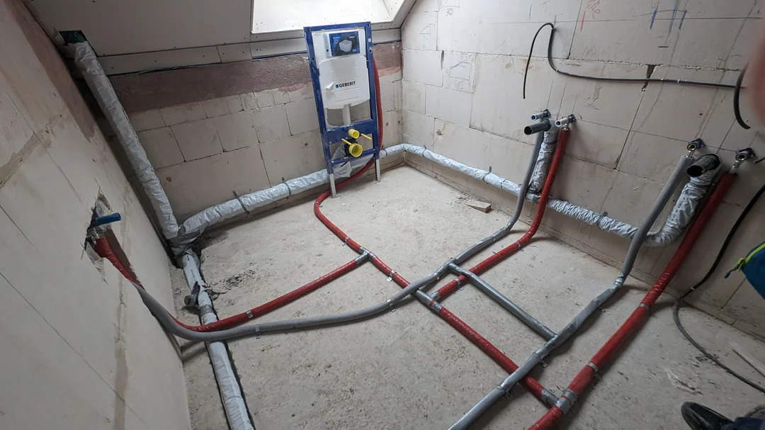 Wnętrze łazienki w trakcie remontu z widoczną instalacją ogrzewania podłogowego z rurami w kolorze czerwonym i szarym, izolowanymi rurami oraz stelażem podtynkowym WC marki Geberit. Ściany surowe...