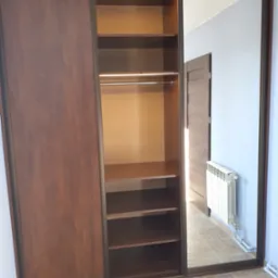 Brązowa szafa wnękowa z przesuwnymi drzwiami, jedna strona z lustrem odbijającym fragment pokoju z grzejnikiem i drzwiami.