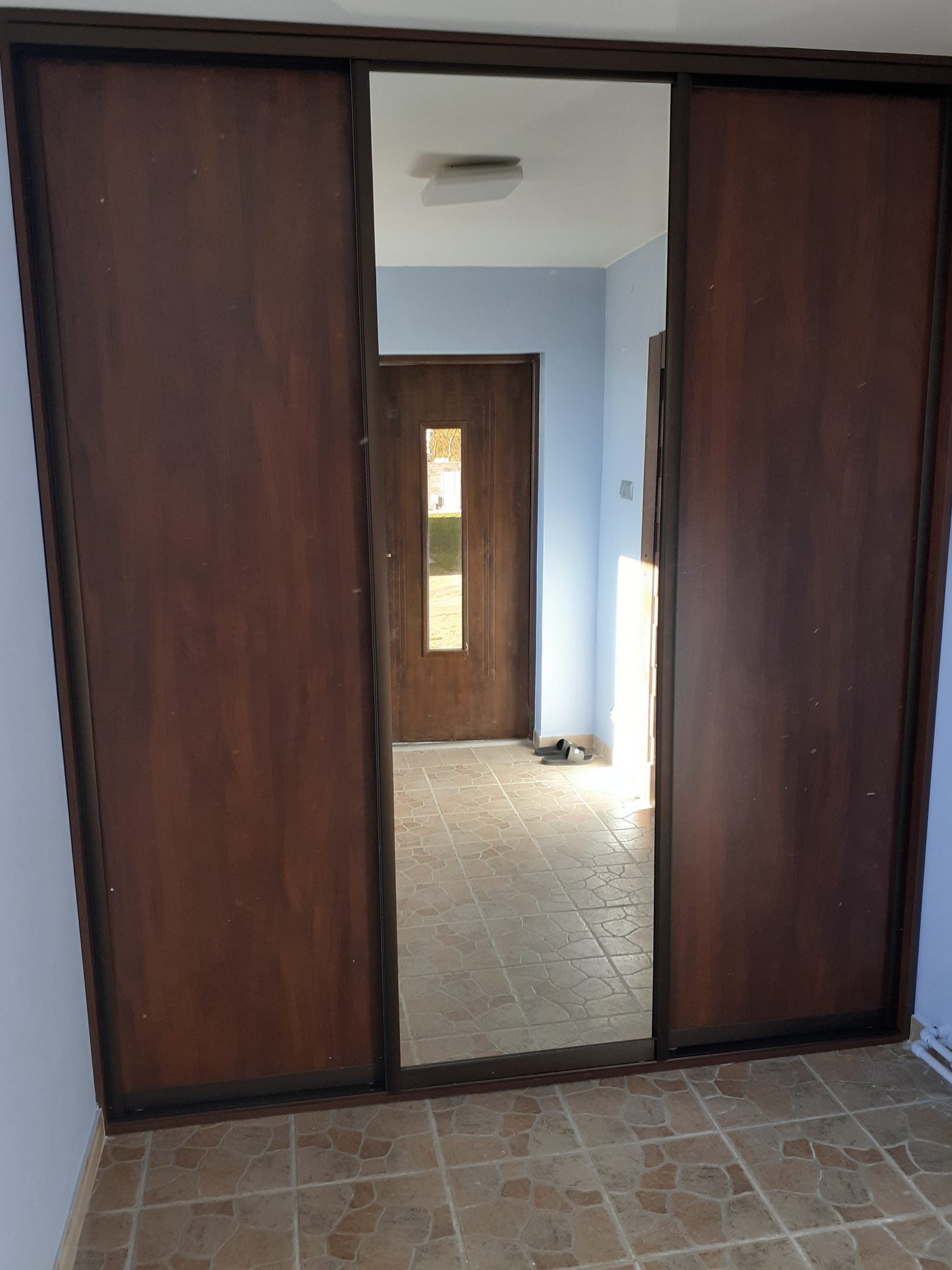 Szafa wnękowa z przesuwnymi drzwiami z ciemnego drewna i lustrem, widoczna w przedpokoju z beżową podłogą i niebieskimi ścianami.