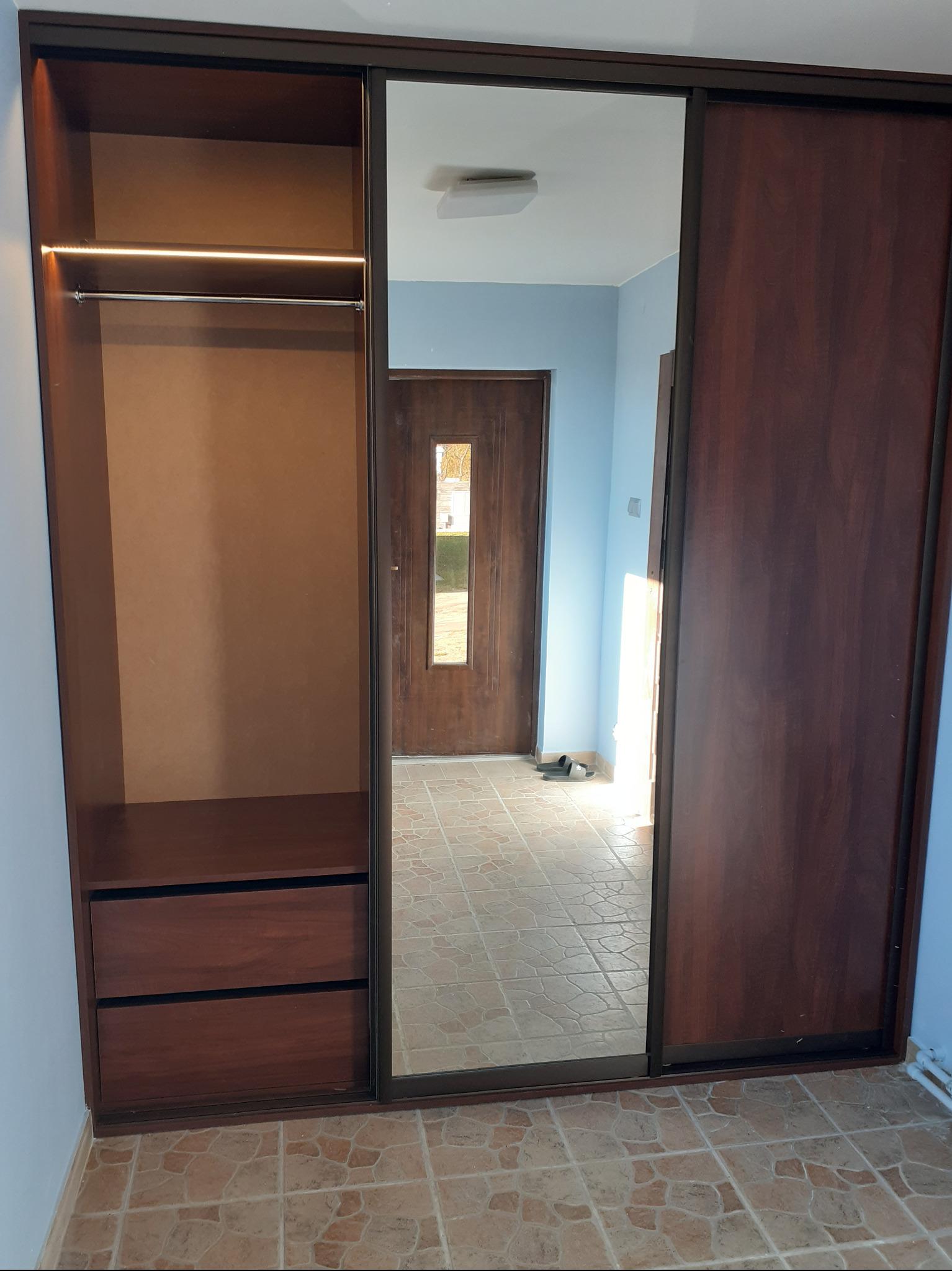 Wnętrze szafy wnękowej z przesuwnymi drzwiami z lustrem, widoczne półki, szuflady i drążek na ubrania z oświetleniem LED, odbicie w lustrze ukazuje fragment przedpokoju z brązowymi drzwiami.