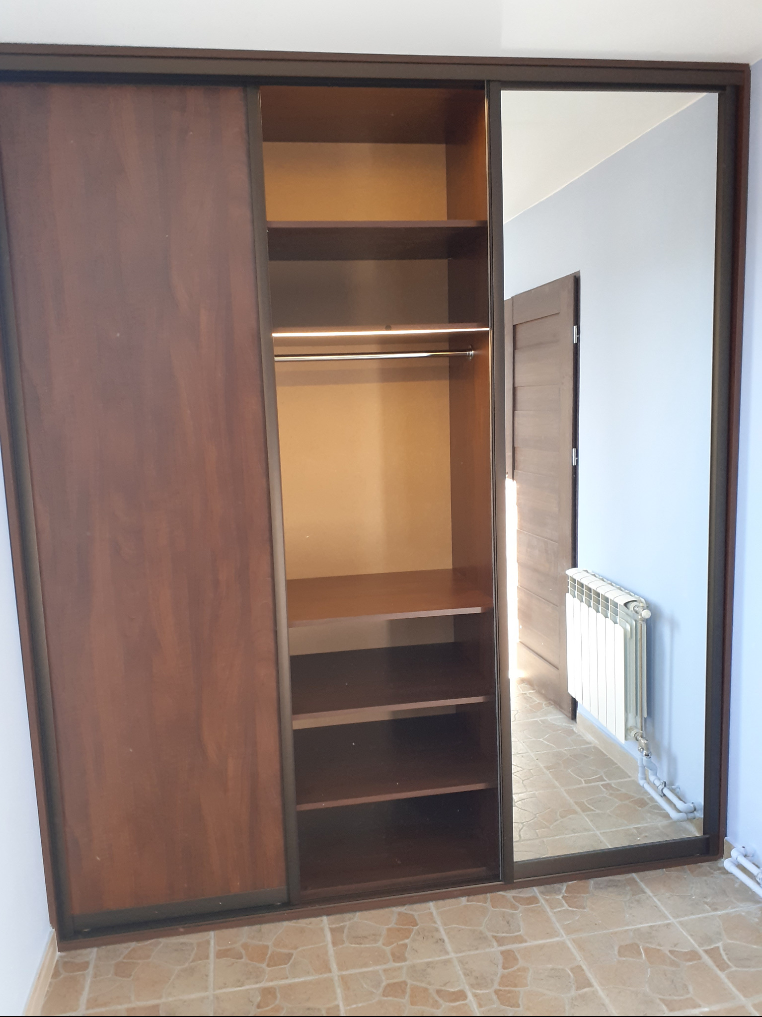 Brązowa szafa wnękowa z przesuwnymi drzwiami, jedna strona z lustrem odbijającym fragment pokoju z grzejnikiem i drzwiami.