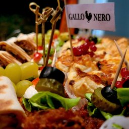 Gallo-nero Katarzyna Stawska - Dekoracyjnie ułożone przekąski na paterze: tortille, winogrona, koreczki z oliwką i ogórkiem, mini tartaletki z czerwoną porzeczką, flaga z logo 'Gallo Nero'.