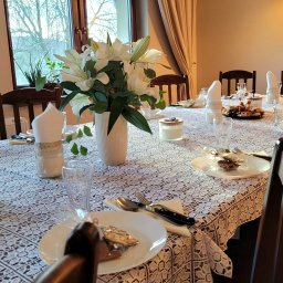 Gallo-nero Katarzyna Stawska - Uroczysty stół nakryty białym obrusem z koronkowym wzorem, z białymi liliami w wazonie, szklankami, serwetkami w słoikach, talerzami z drobnymi przekąskami i upominkami dla gości