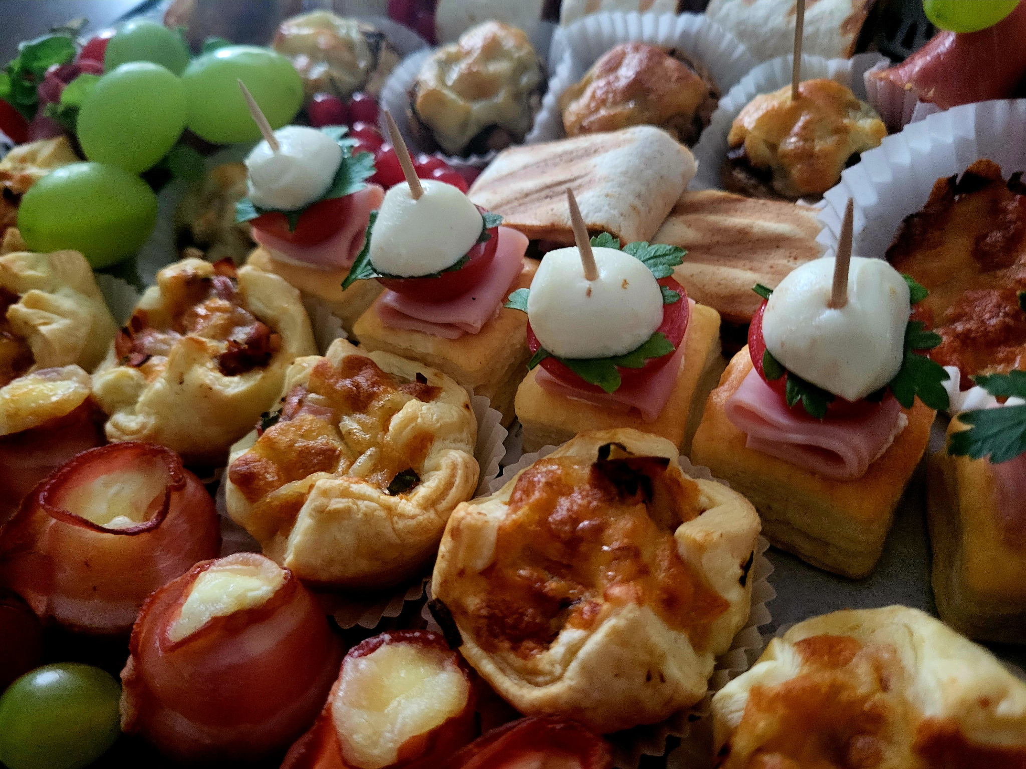 Patera z różnorodnymi przekąskami: mini tortille z nadzieniem, saszetki z ciasta francuskiego z suszonymi pomidorami, mozzarella, natką pietruszki, szynką, pieczarki faszerowane, oraz winogrona...