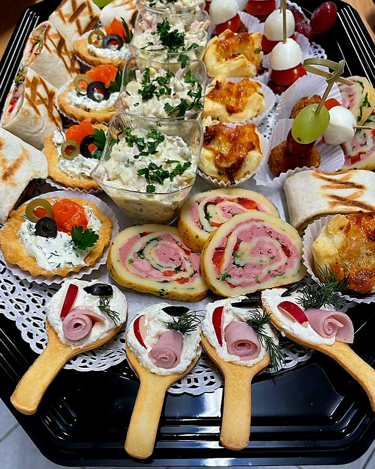 Czarny prostokątny półmisek z różnorodnymi przekąskami: rolada serowa z szynką, babeczki z łososiem, mini tartaletki z kremowym serem i oliwkami, koreczki z mozzarelli i pomidorków, sałatki...