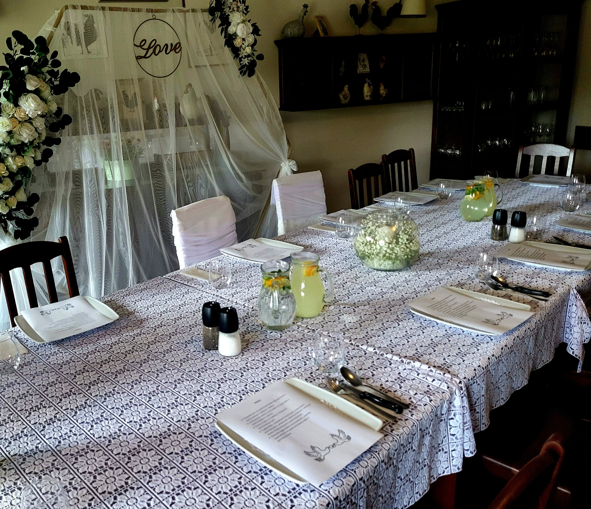 Uroczysty stół nakryty białym koronkowym obrusem, z dekoracjami kwiatowymi, dzbankami z lemoniadą, solniczkami i pieprzniczkami oraz menu z motywem gołębi, przygotowany na przyjęcie.