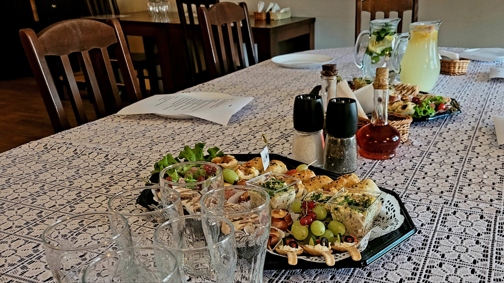 Stół nakryty białym obrusem w koronkowy wzór, na nim taca z przekąskami, szklanki, dzbanki z napojami, solniczka i pieprzniczka, oraz kartka z menu.
