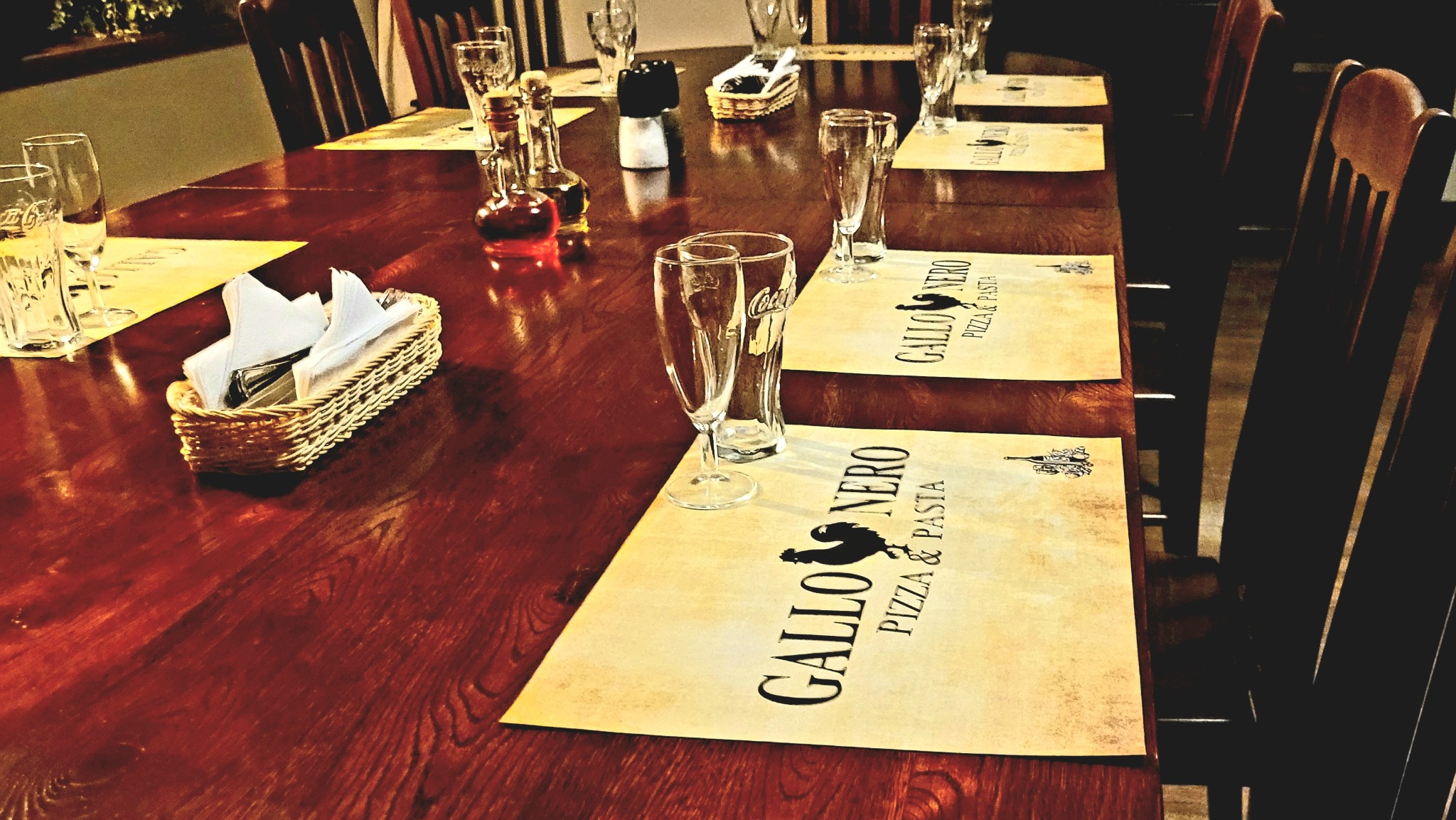 Długi, drewniany stół nakryty dla gości w restauracji: szklanki, serwetki w koszyczku, butelki z przyprawami oraz podkładki z logo 'Gallo Nero Pizza & Pasta'.