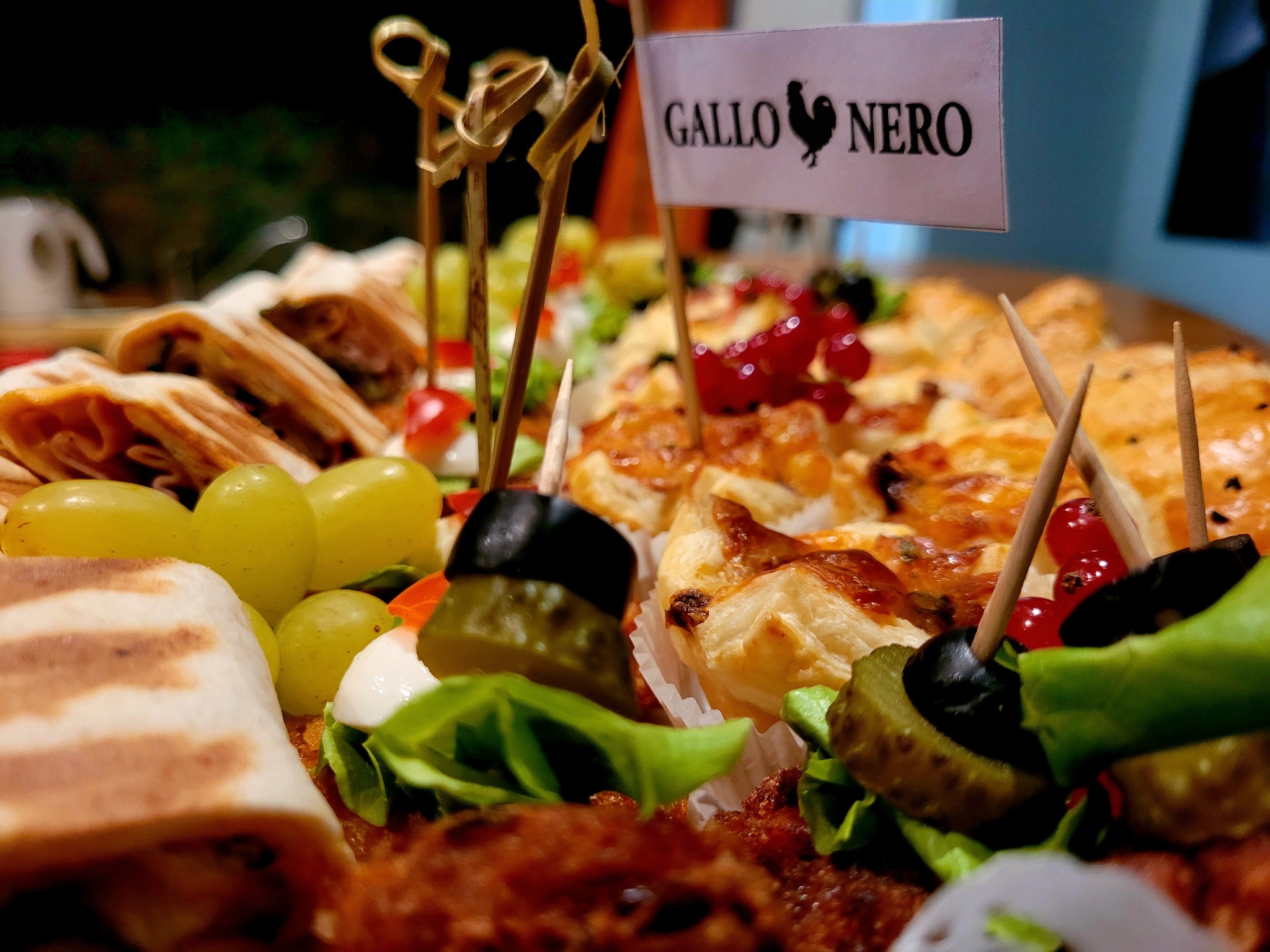 Dekoracyjnie ułożone przekąski na paterze: tortille, winogrona, koreczki z oliwką i ogórkiem, mini tartaletki z czerwoną porzeczką, flaga z logo 'Gallo Nero'.
