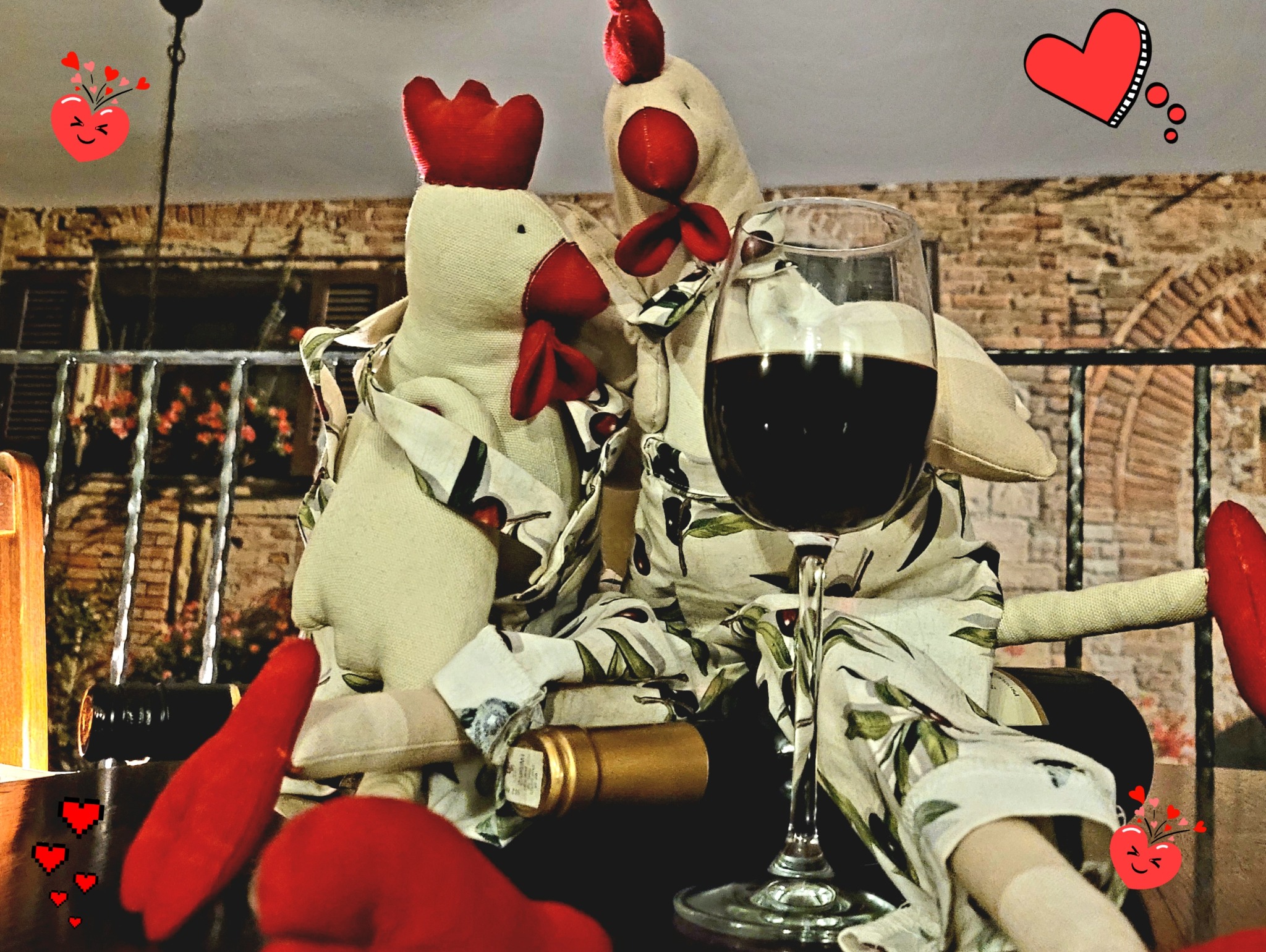 Dwa pluszowe koguty w koszulach w kwiaty siedzą obok kieliszka czerwonego wina i butelki, na tle imitacji ceglanej ściany i balkonu z kwiatami. Dodane grafiki z sercami.