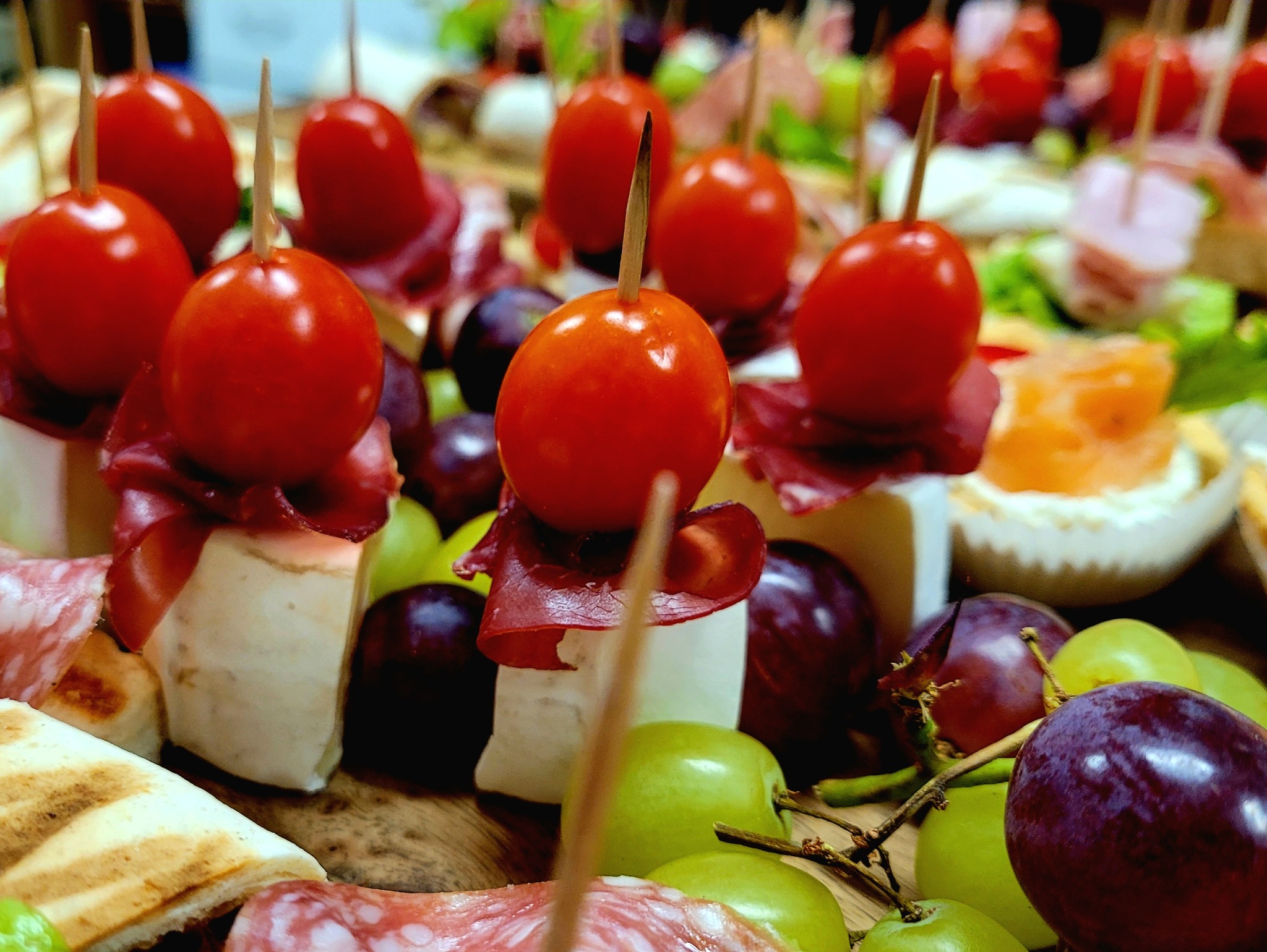 Zbliżenie na deskę z przekąskami: koreczki z pomidorków cherry, szynki i sera, ciemne i zielone winogrona, salami oraz mini tartaletki z łososiem, ułożone na drewnianej desce.
