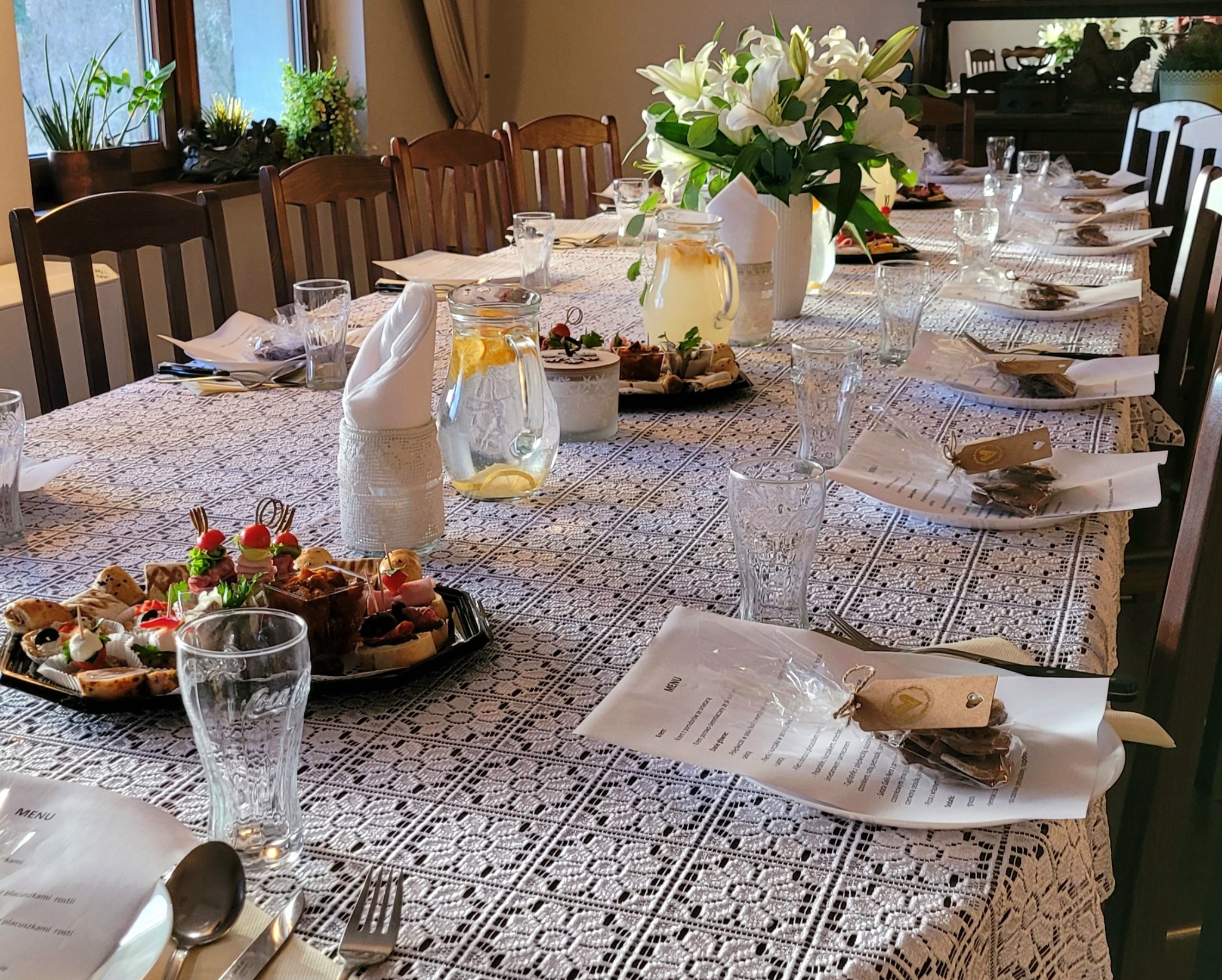 Uroczysty stół nakryty koronkowym obrusem, z przygotowanymi porcjami przekąsek na talerzach, dzbankami z lemoniadą, bukietem białych lilii i menu dla gości.