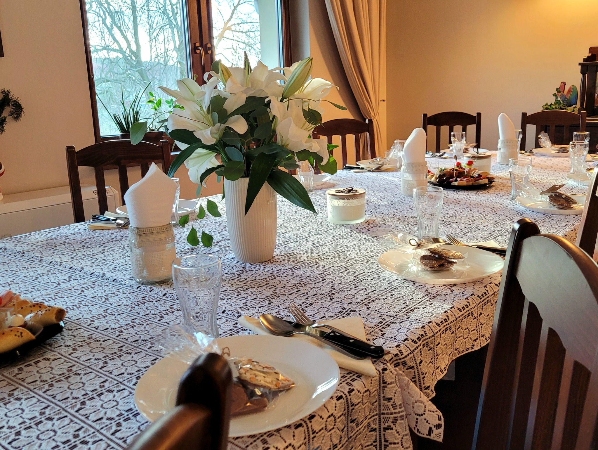 Uroczysty stół nakryty białym obrusem z koronkowym wzorem, z białymi liliami w wazonie, szklankami, serwetkami w słoikach, talerzami z drobnymi przekąskami i upominkami dla gości