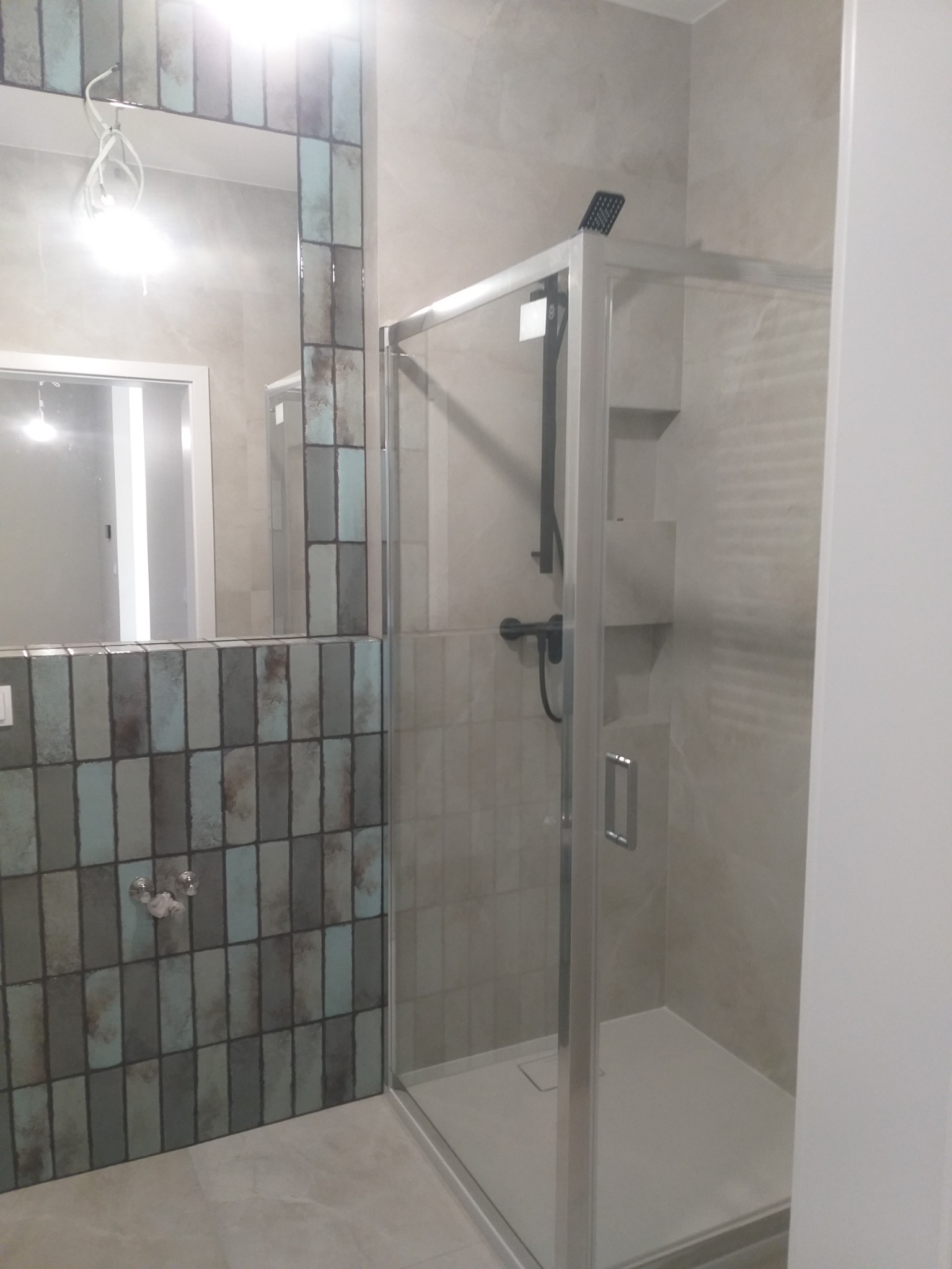 Szklana kabina prysznicowa z czarną armaturą, zamontowana w łazience z płytkami w odcieniach szarości i błękitu, widoczny fragment lustra z dekoracyjną obudową z płytek.