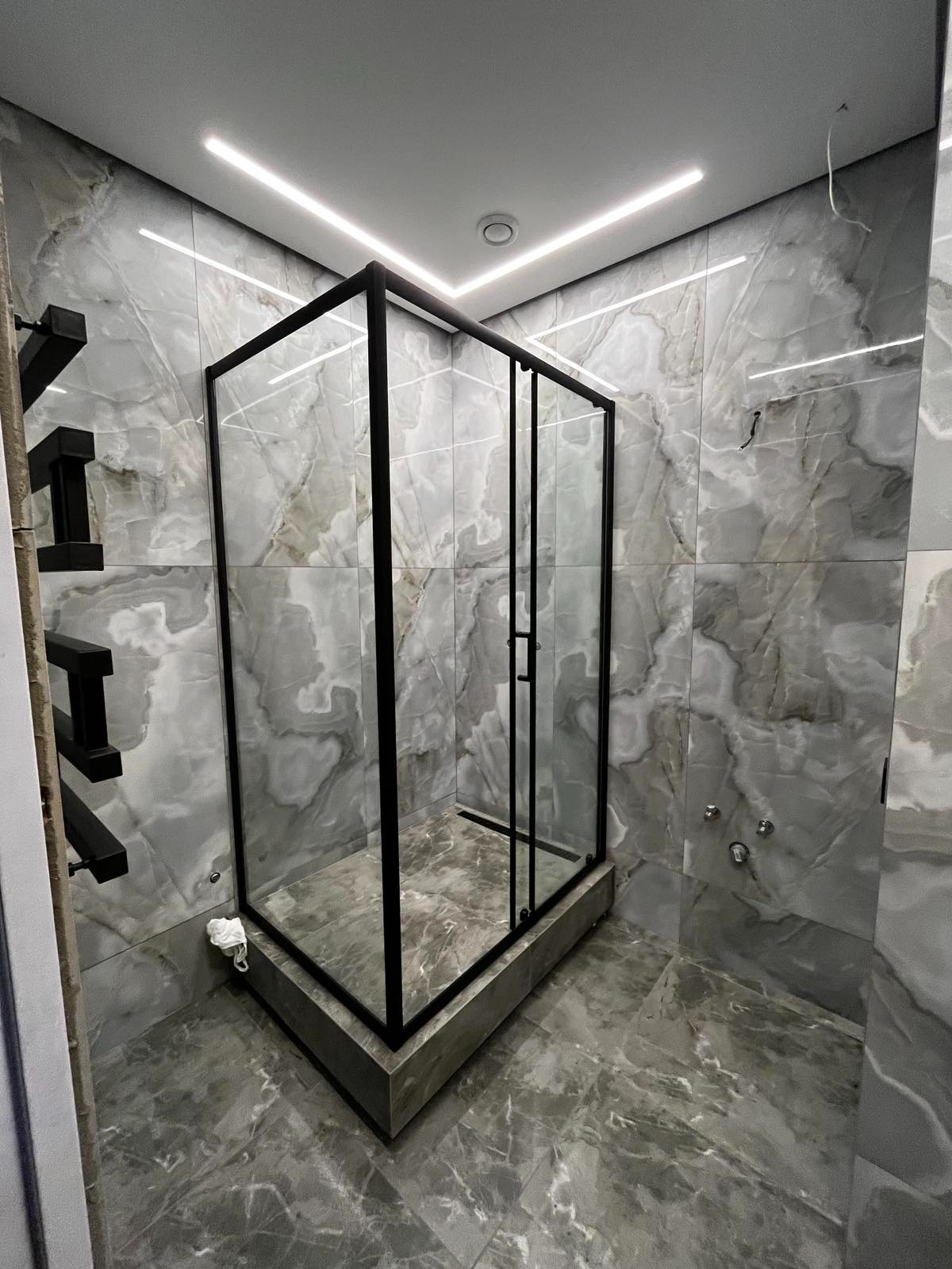Nowoczesna kabina prysznicowa z czarnymi profilami i szklanymi ściankami, umieszczona w łazience wykończonej szarym marmurem, z oświetleniem LED w suficie.