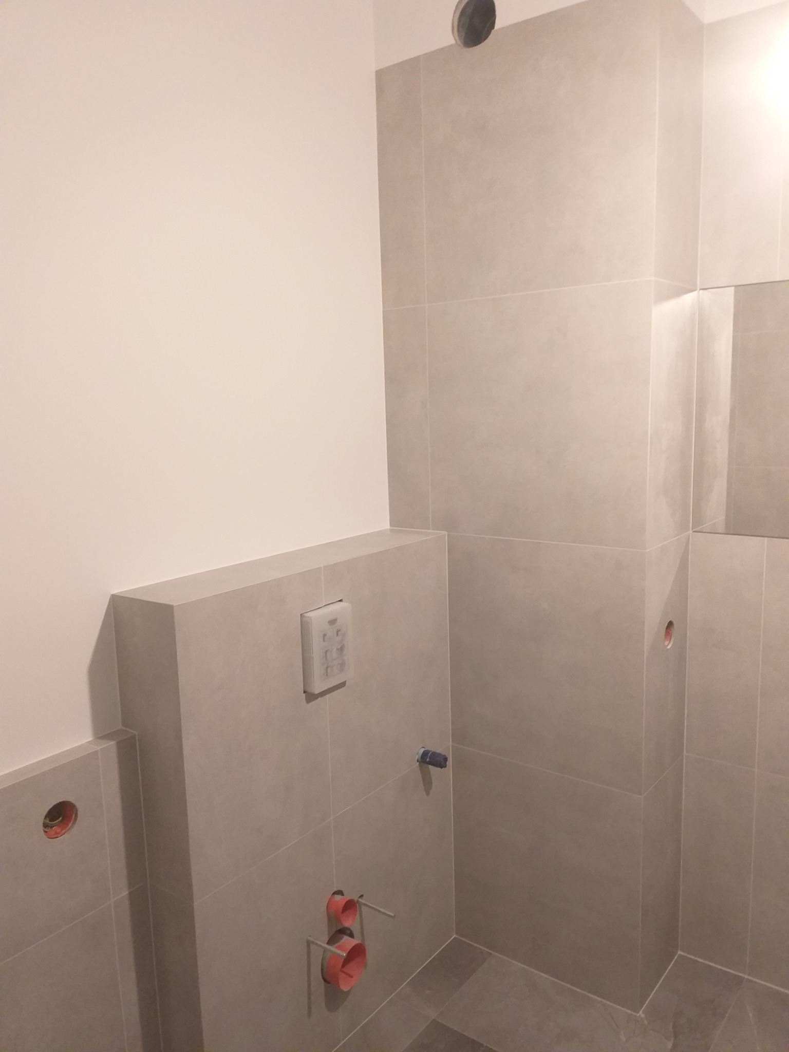 Łazienka w trakcie wykańczania: szare płytki na ścianach i podłodze, widoczne otwory na instalacje, zamontowany stelaż podtynkowy WC z przyciskiem spłukującym, sufit z otworem wentylacyjnym.