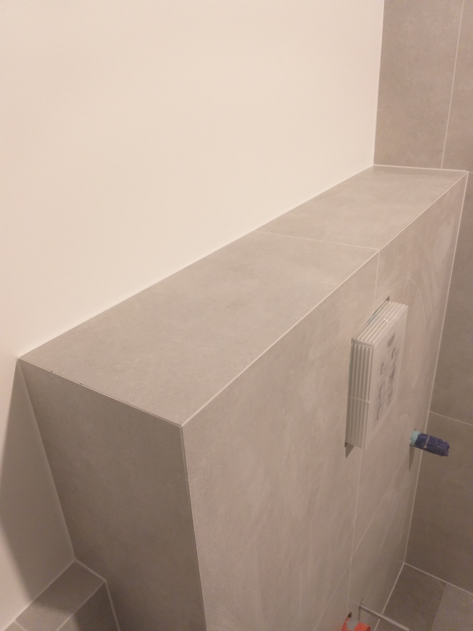 Szara zabudowa stelaża podtynkowego WC z przyciskiem spłukującym, wykończona płytkami ceramicznymi w łazience.