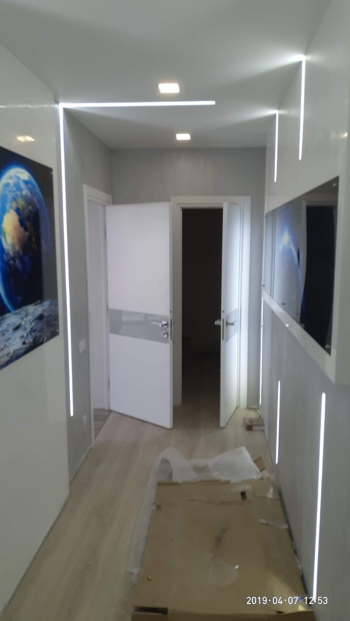 Korytarz z podświetlanymi listwami LED wzdłuż ścian i sufitu, częściowo zabezpieczony kartonem i folią malarską, z widocznymi drzwiami wewnętrznymi.