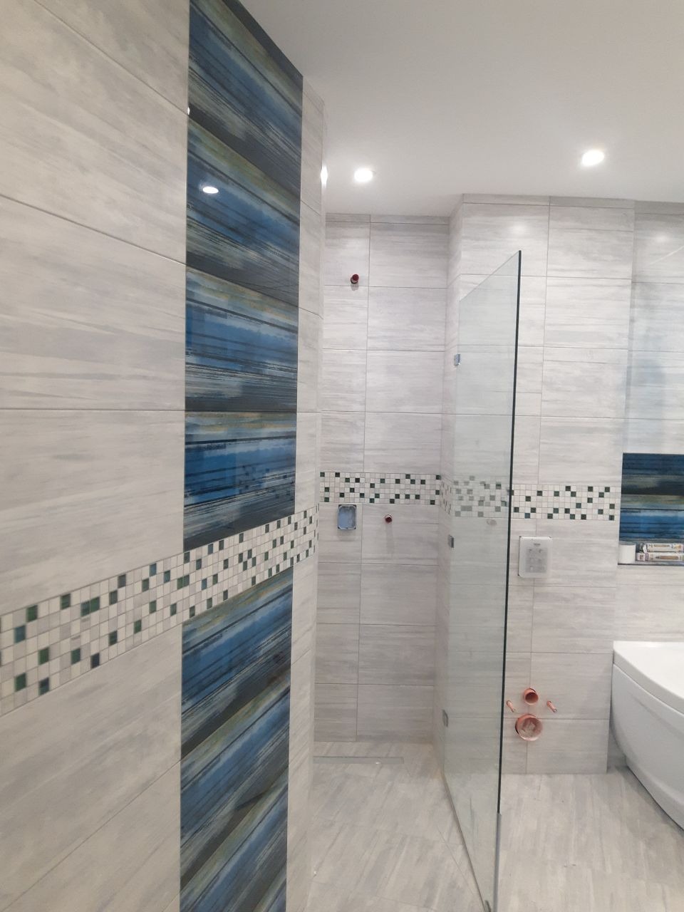 Wnętrze łazienki z jasnymi, podłużnymi płytkami imitującymi drewno, akcentem dekoracyjnym z niebieskich pasów i mozaiki oraz kabiną prysznicową ze szkła.