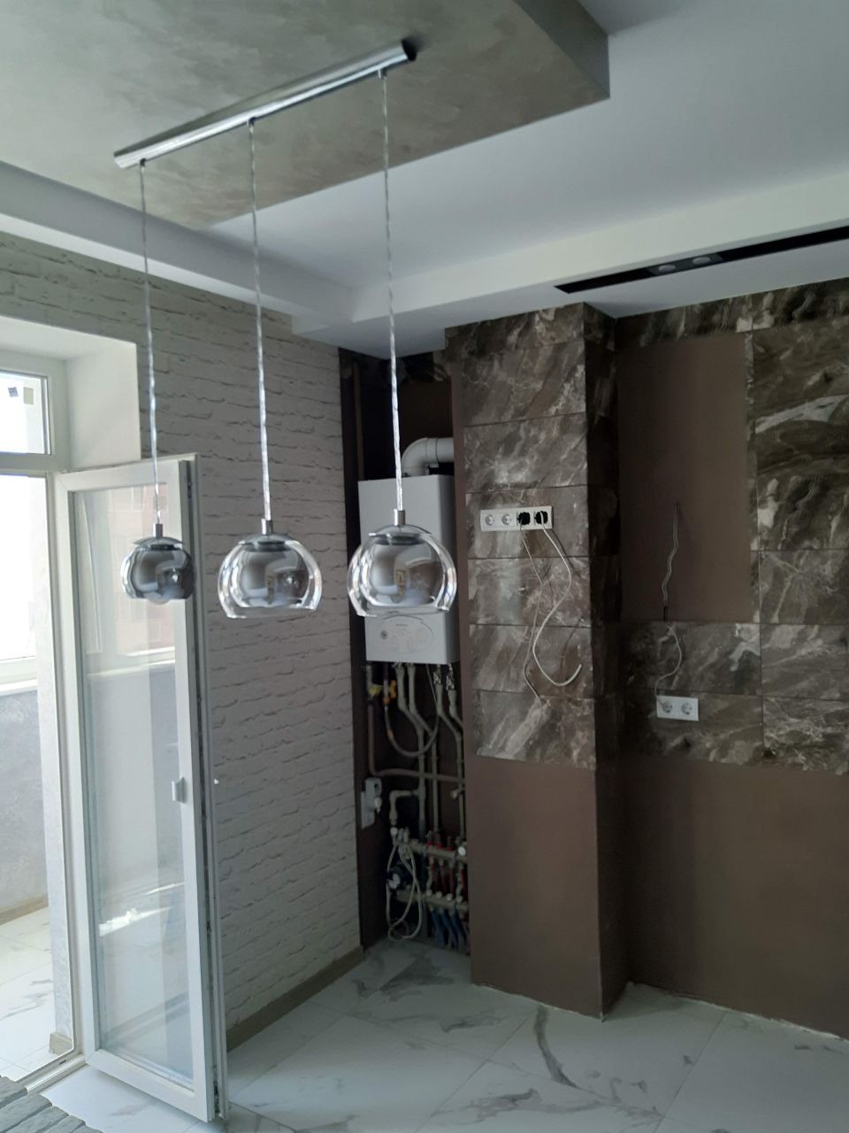 Nowoczesne, szklane lampy wiszące nad balkonem z otwartymi drzwiami, marmurowa okładzina ściany i widoczne instalacje grzewcze.
