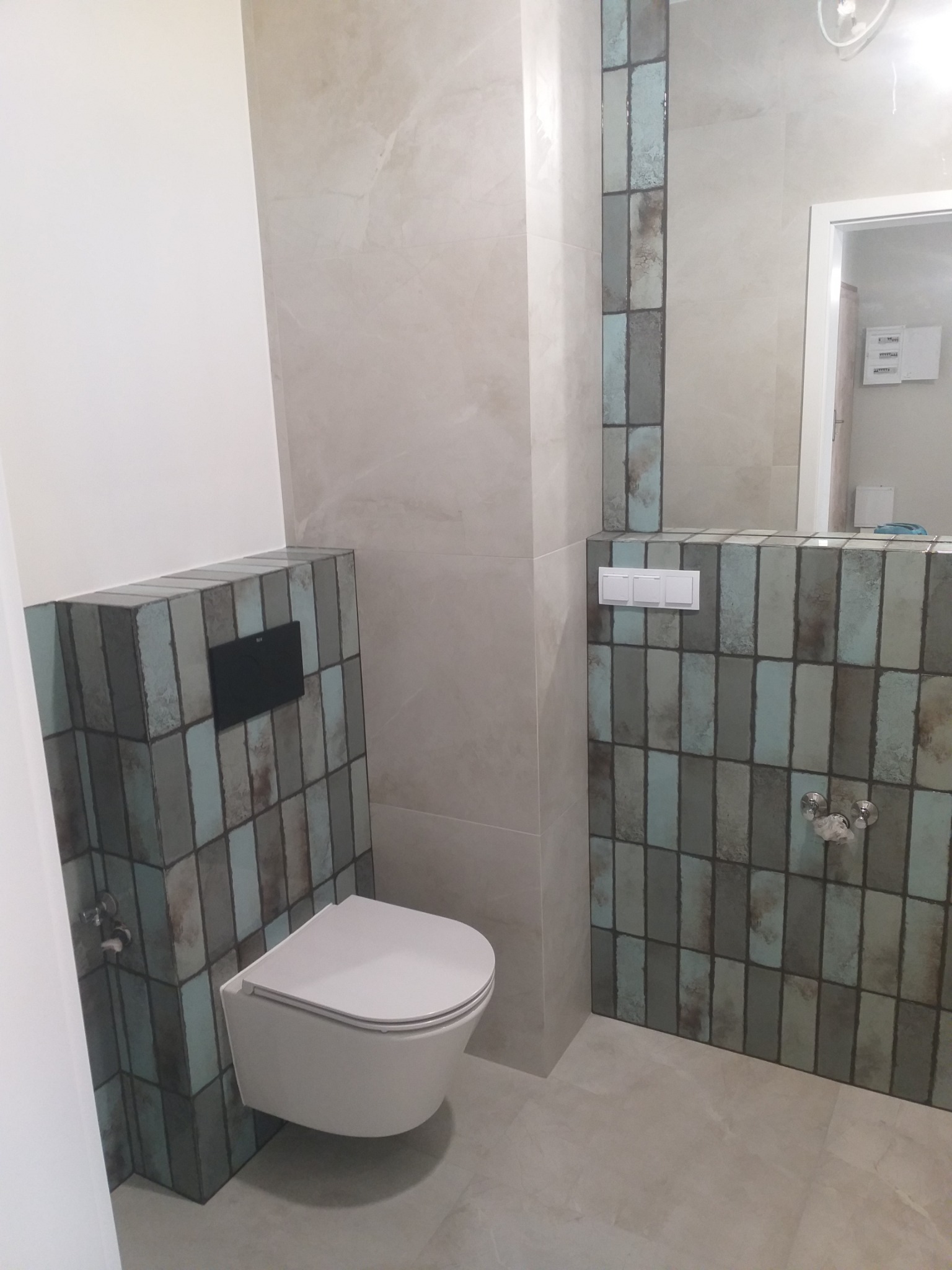 Nowoczesna łazienka z podwieszaną toaletą, obudową z płytek w odcieniach szarości i błękitu, oraz beżowymi płytkami na ścianach i podłodze.